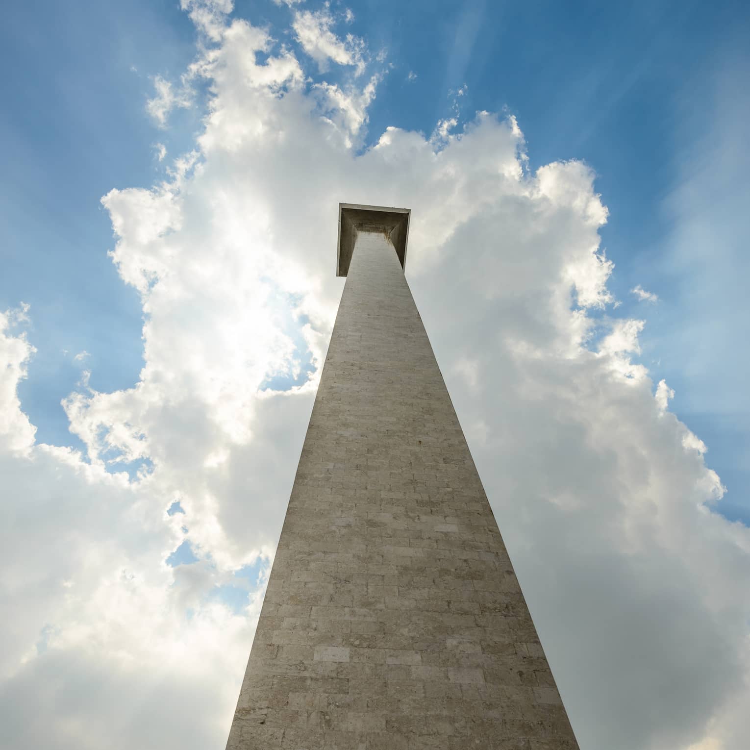 Staring up from the base of the Monas National Monument; a blue sky and the sun peeking through the clouds as a backdrop.