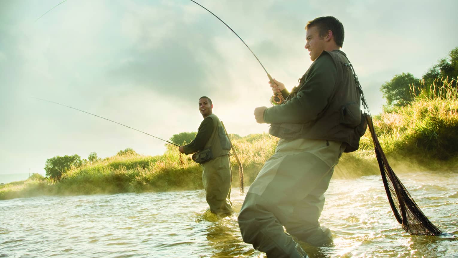 Two people fly fishing in a river, standing knee-deep in the water, surrounded by mist and greenery.