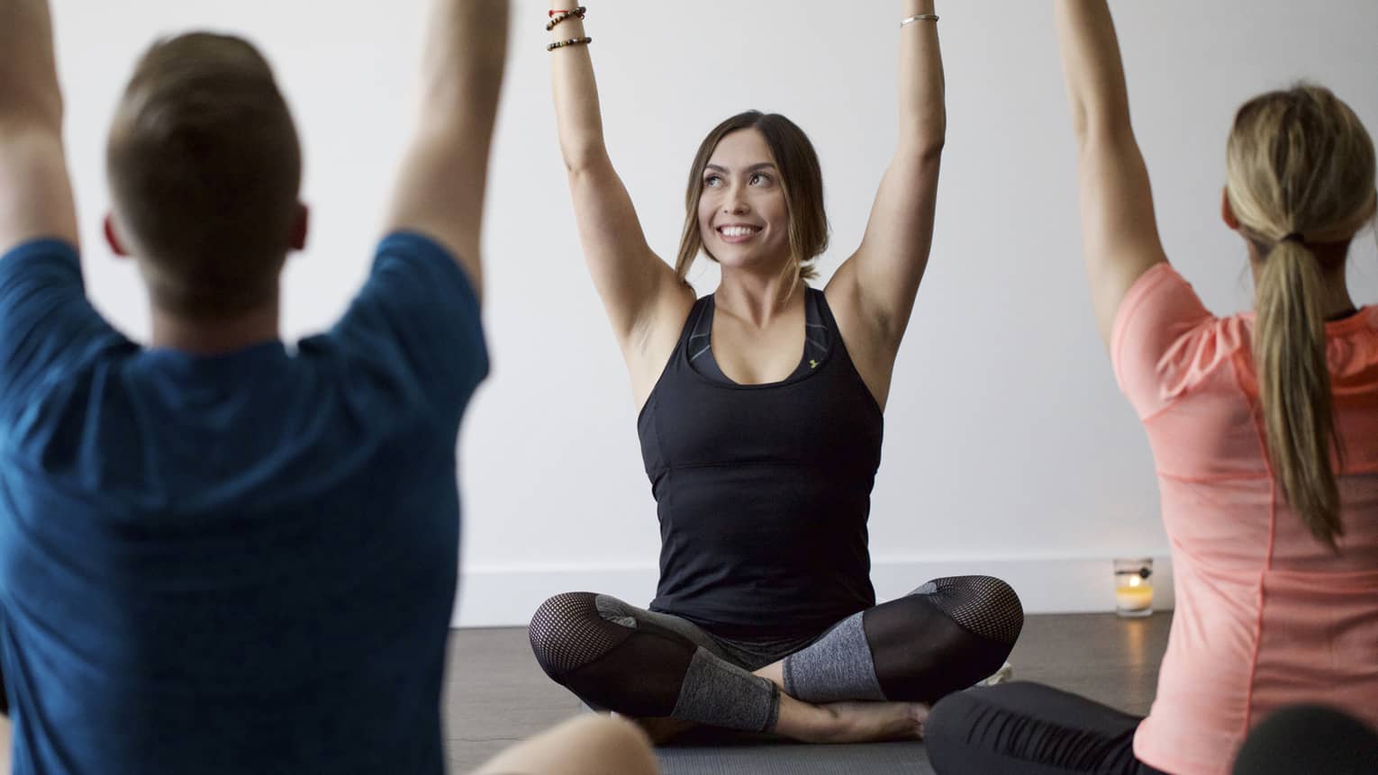 Man, woman sit in front of smiling yoga instructor, cross-legged with arms raised above head