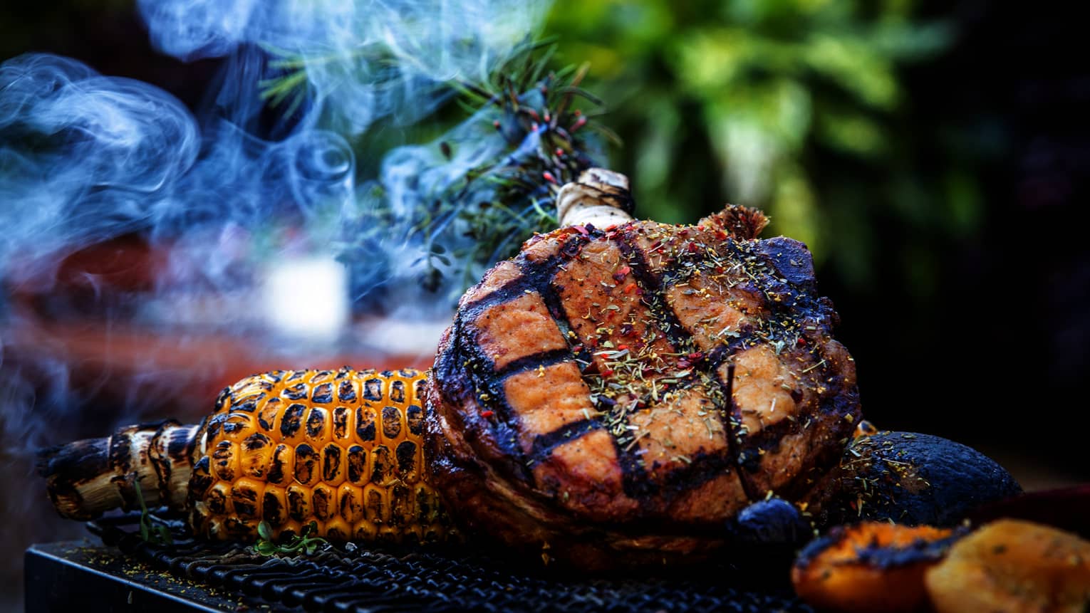 A grill with meat, corn and potatoes cooking on it outside.