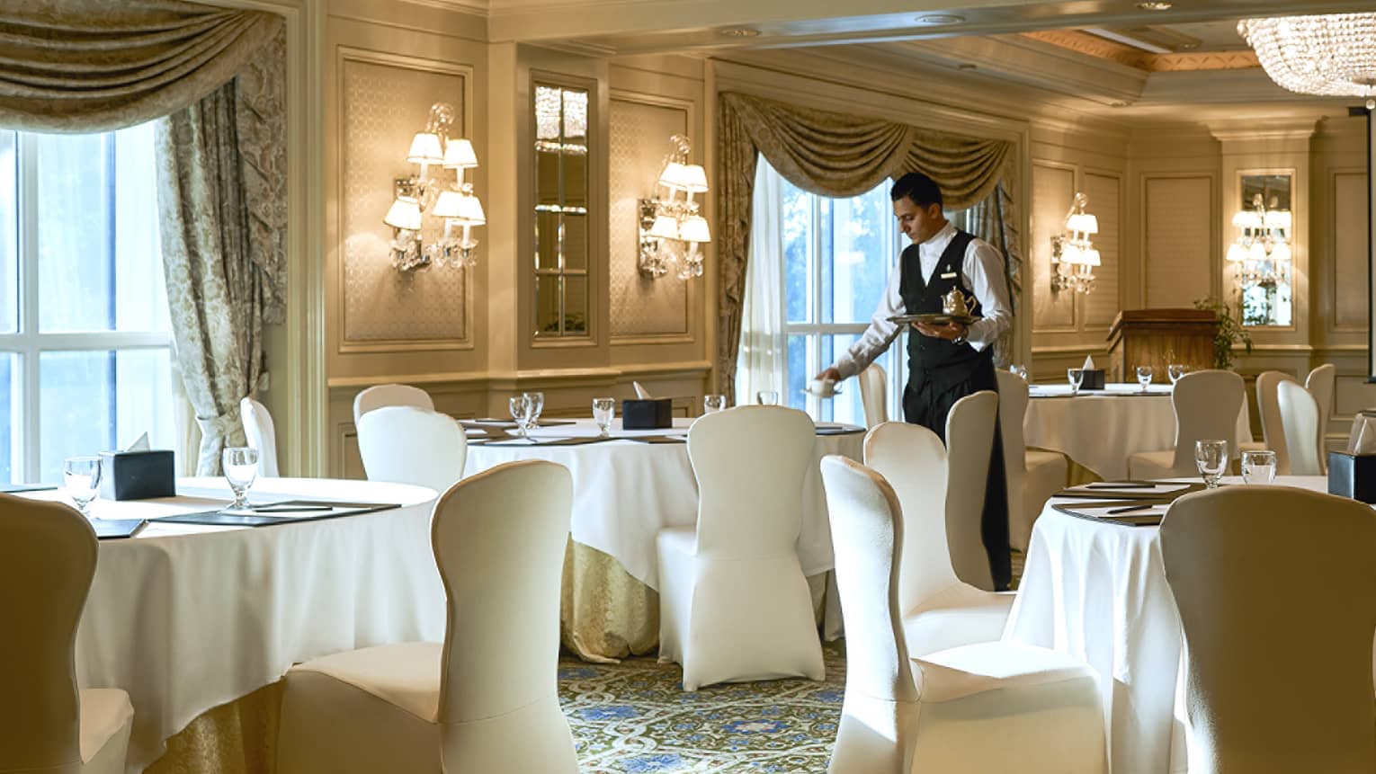 Hotel waitstaff sets small round dining tables with white linens near windows 