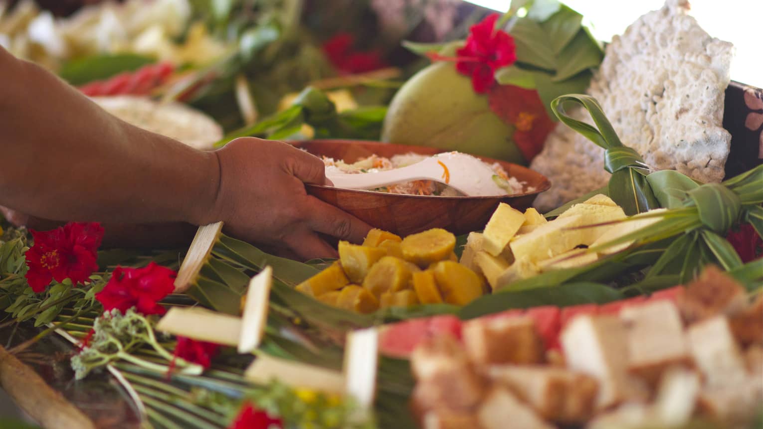 Hand grasps wooden bowl in display of fresh tropical fruit adorned with red flowers