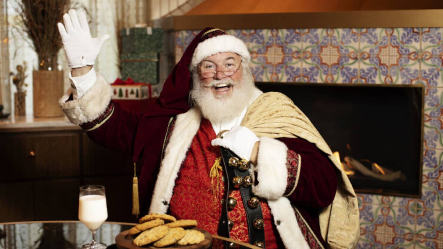 Santa Claus sitting at a table with a plate of cookies and glass of milk, and a fireplace in the background.
