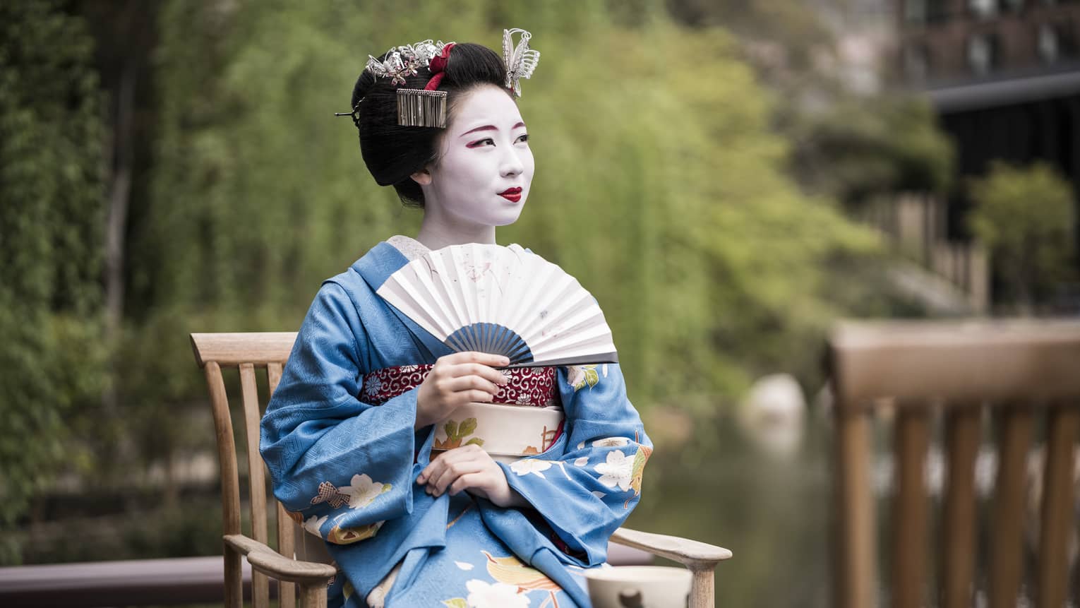 Geiko, a traditional Japanese entertainer, dressed in kimono and holding fan, sitting with cup of tea in garden