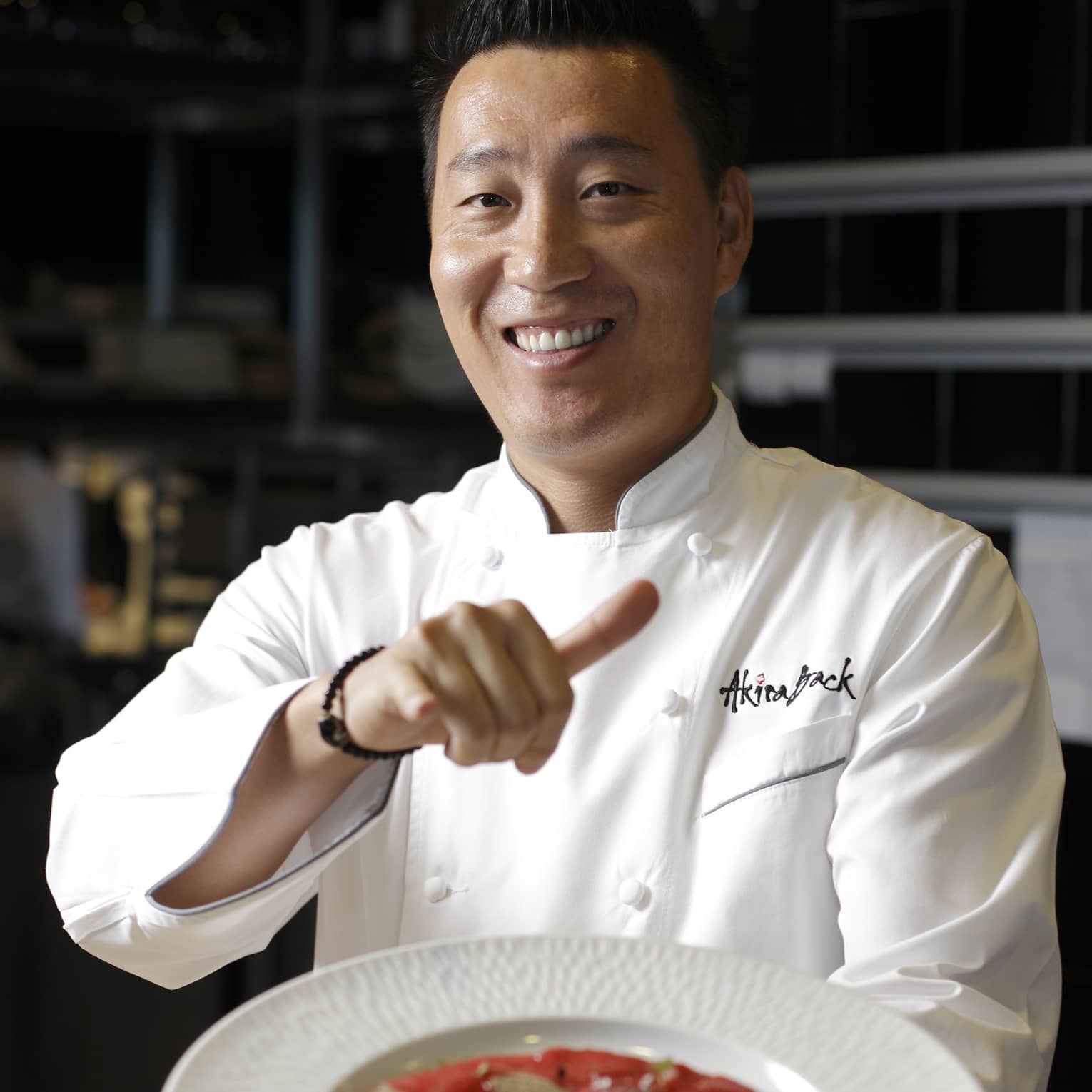 Smiling chef presents a signature dish of tuna pizza garnished with slices of black truffle in a wide, white bowl.