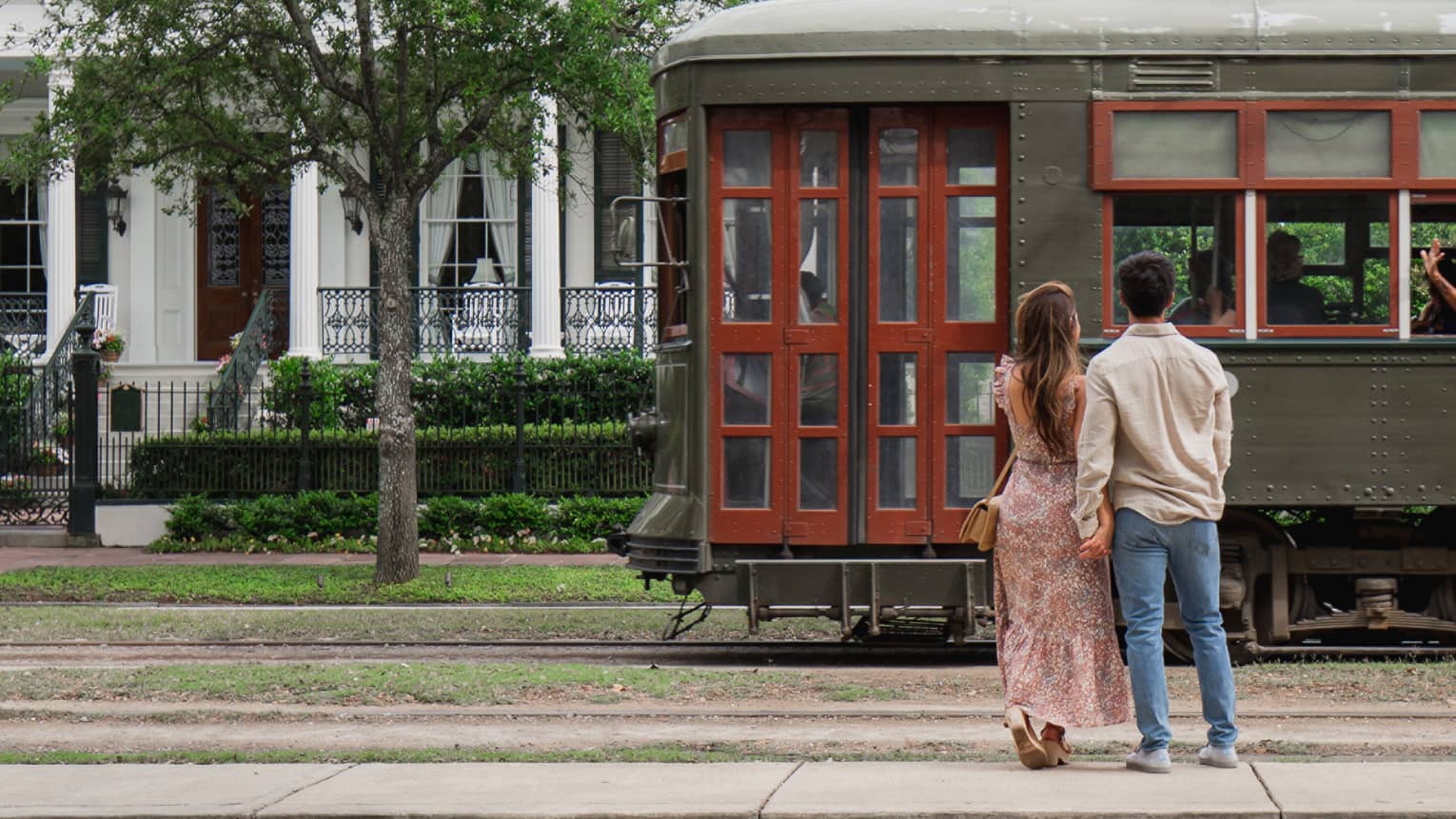 Woman and man hold hands in front of old-fashioned streetcar on New Orleans street, white house in background