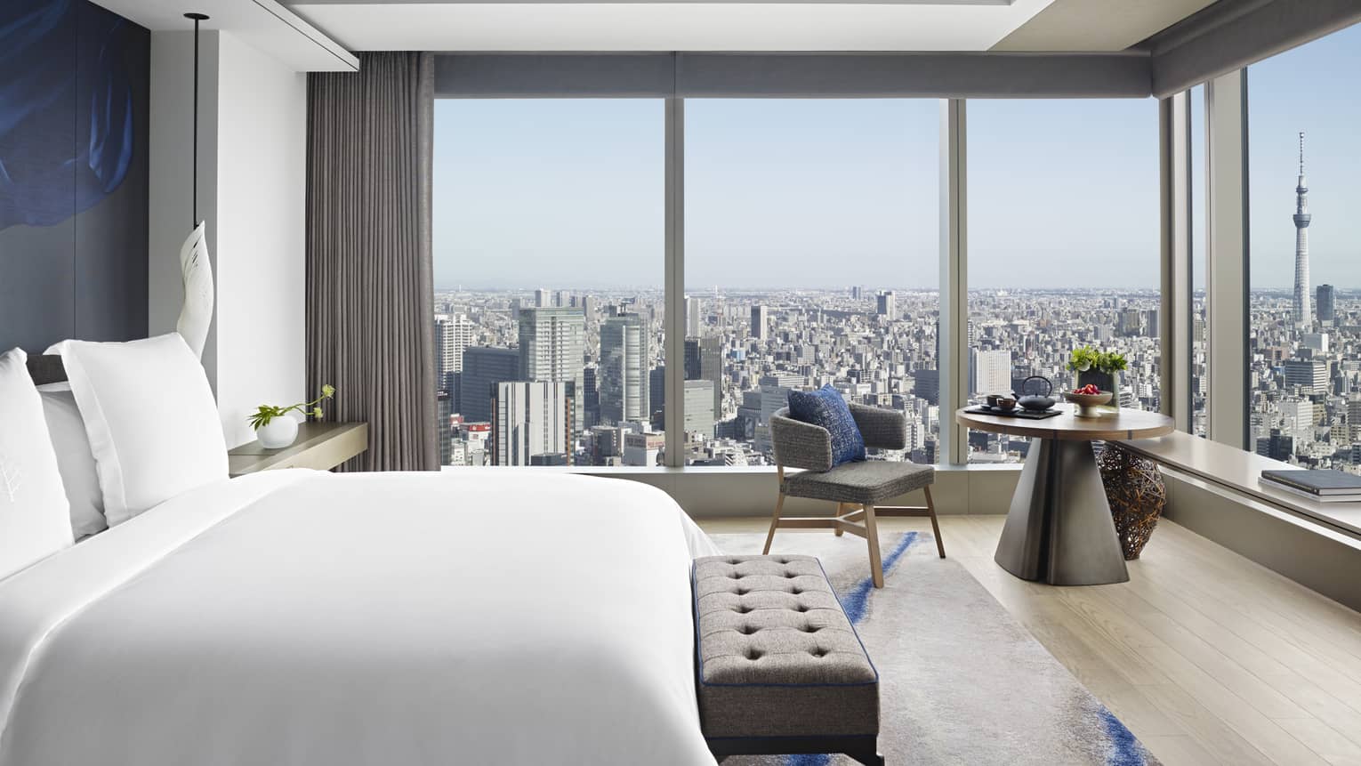 Guest room with king bed, tufted bench, round table with armchair and floor-to-ceiling windows offering city views