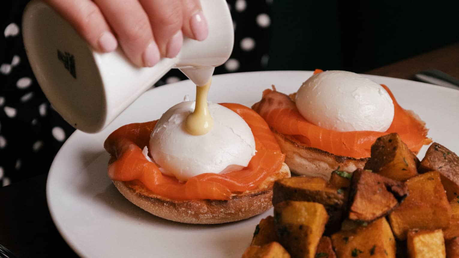 Sauce being poured over two poached eggs with smoked salmon on English muffins, served alongside roasted potatoes.