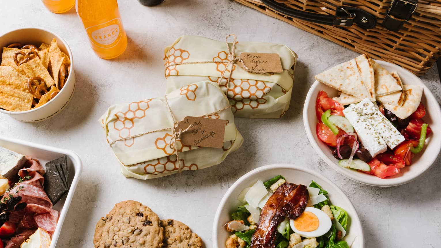 An assortment of picnic food including salad, cookies, sandwiches, fruit, and sliced meat next to a brown wood basket.
