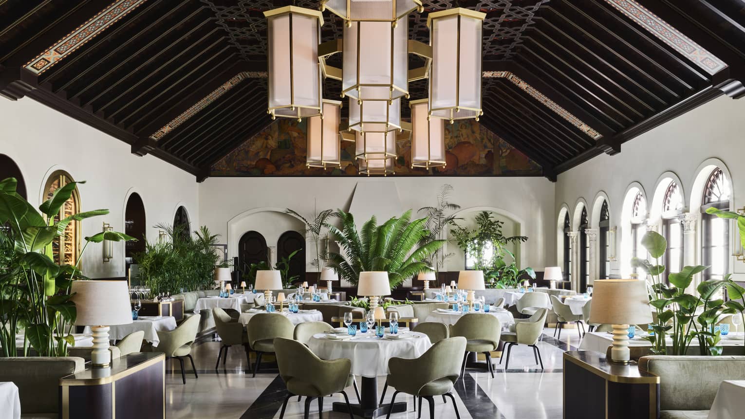 A sophisticated dining room with high, dark wood-beamed ceilings, large hanging lantern chandeliers, green upholstered chairs, white tablecloths and indoor plants.