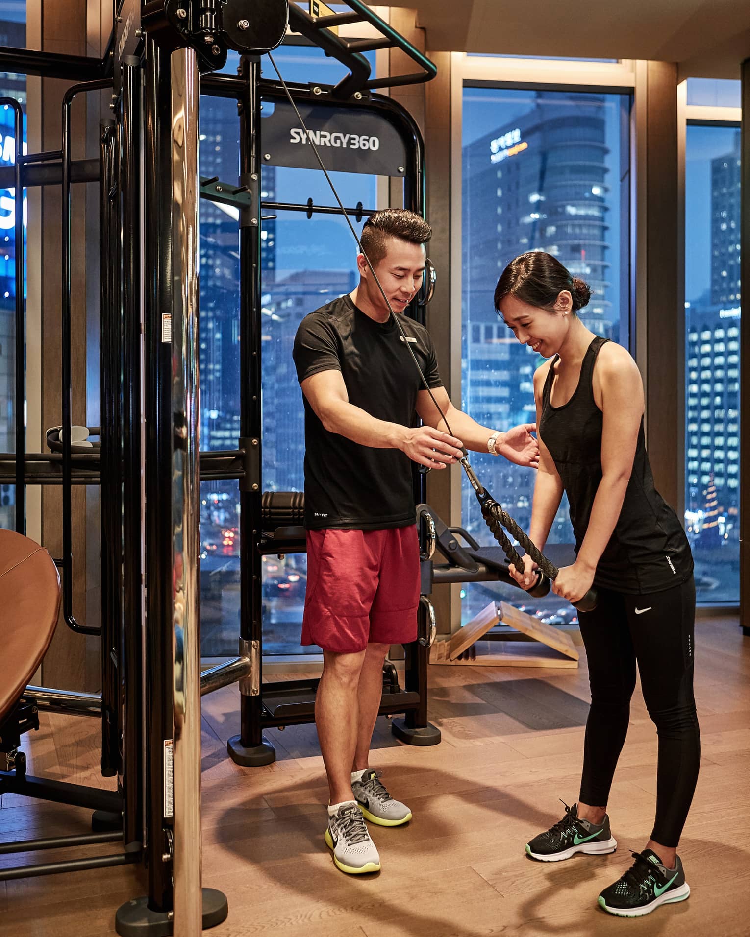 With skyscrapers out the window beyond, a trainer guides a guest as she performs a triceps pushdown exercise on a gym machine.