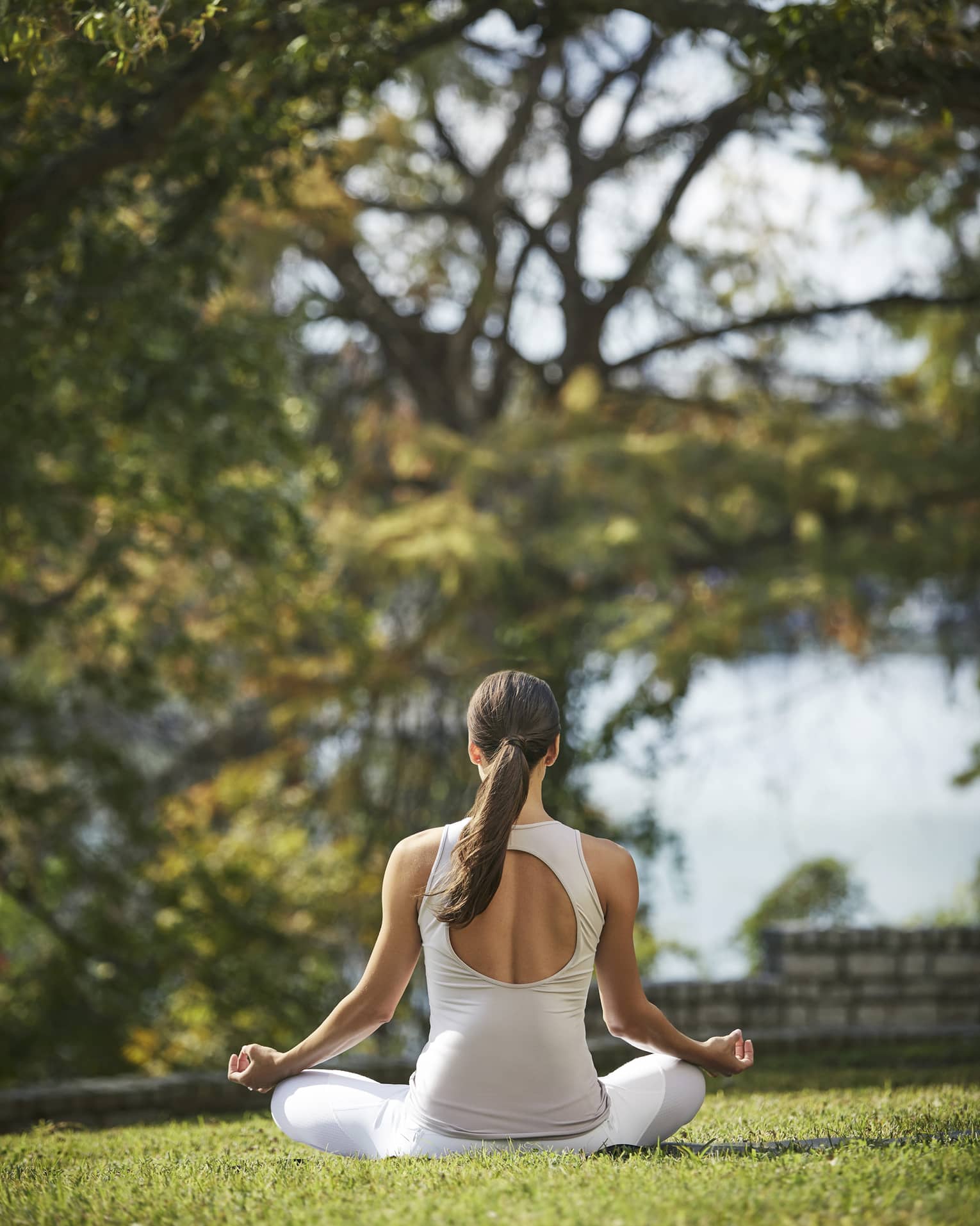 Rear view of a guest in white athletic wear meditating cross-legged with forearms rested on knees on a sunny patch of grass.