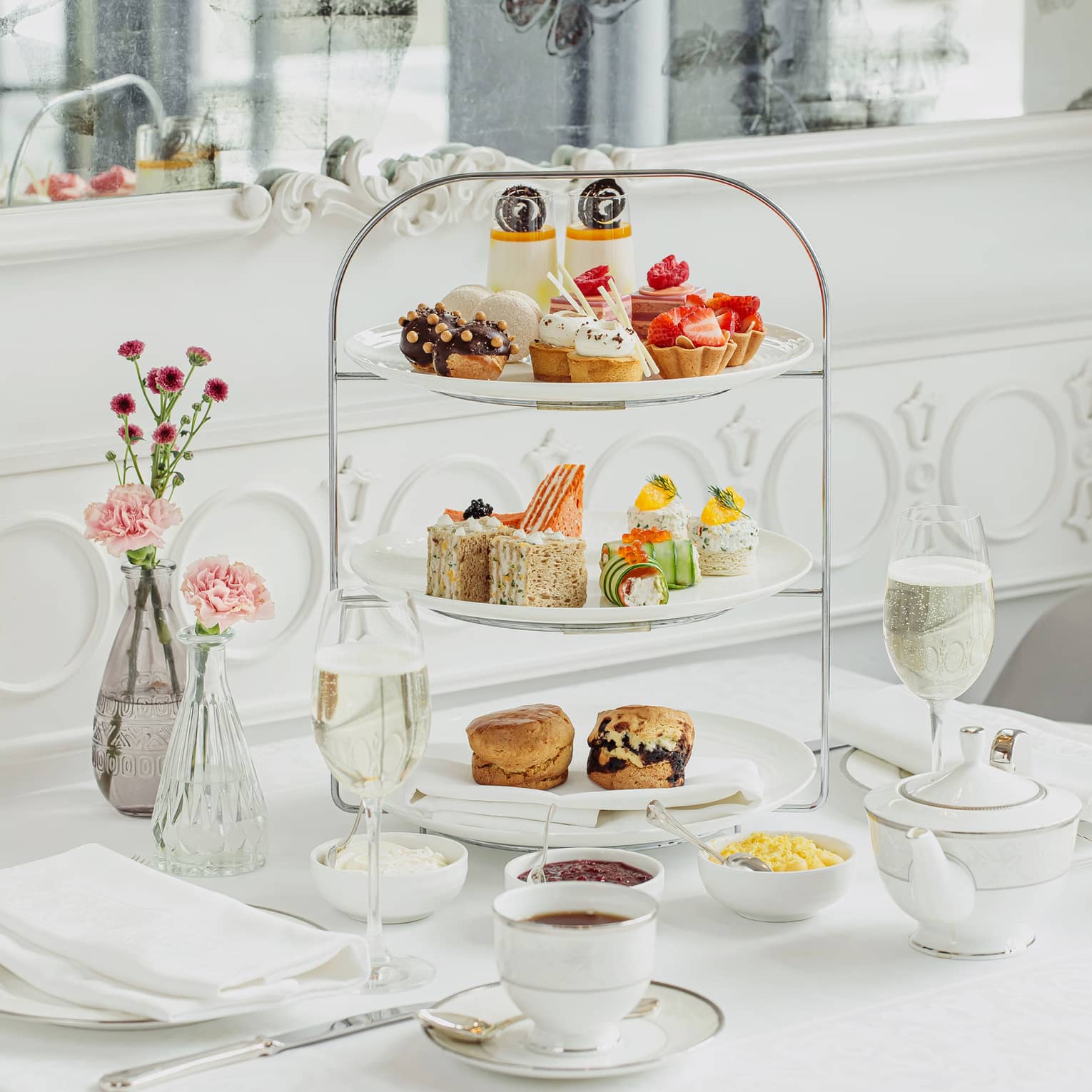 Side dishes, drinks and flowers surround a delicately tiered cake stand holding colourful selections of petite baked goods.
