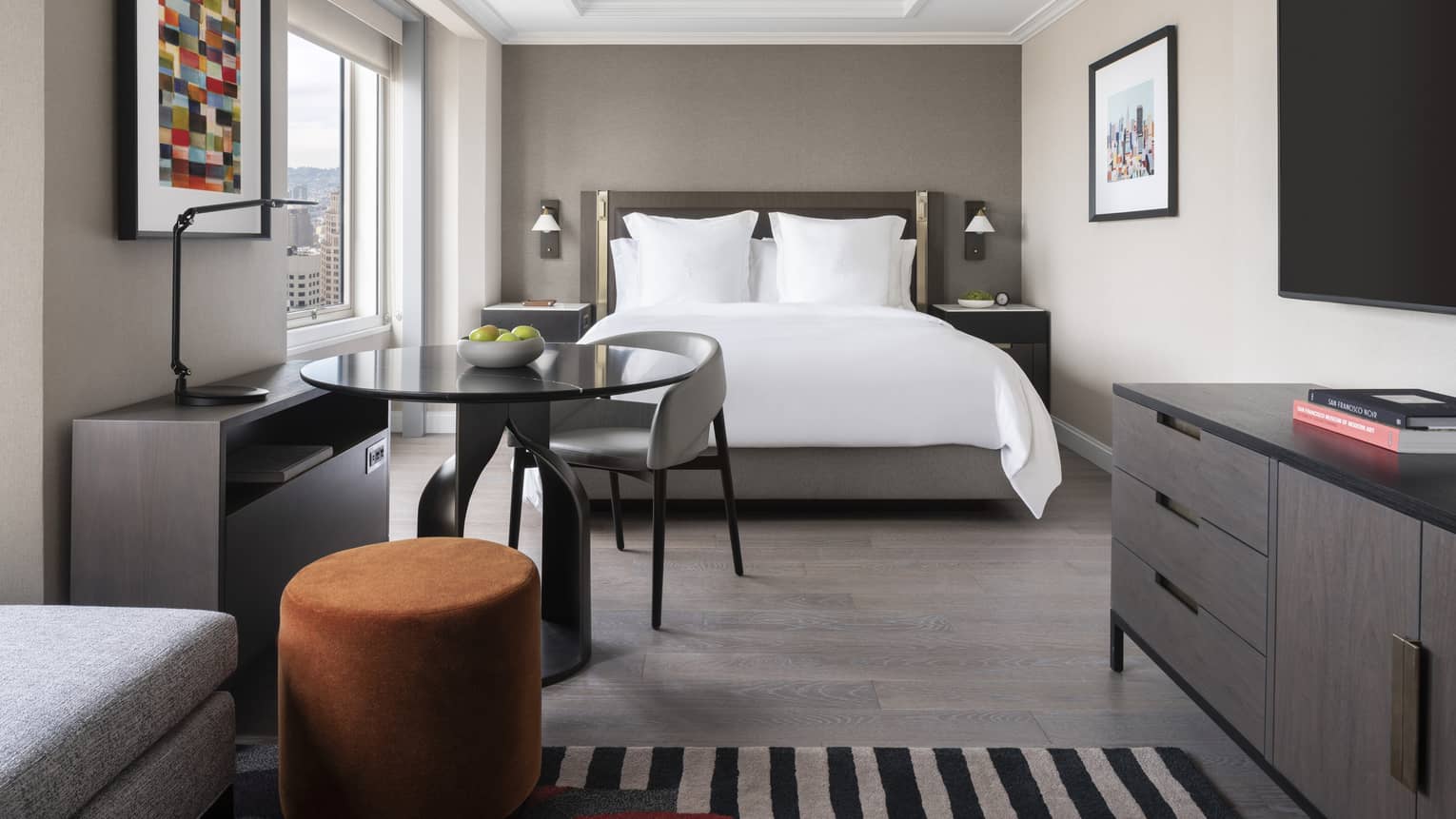 A hotel room with a large bed with white sheets, a desk, a tv, a chair and table.