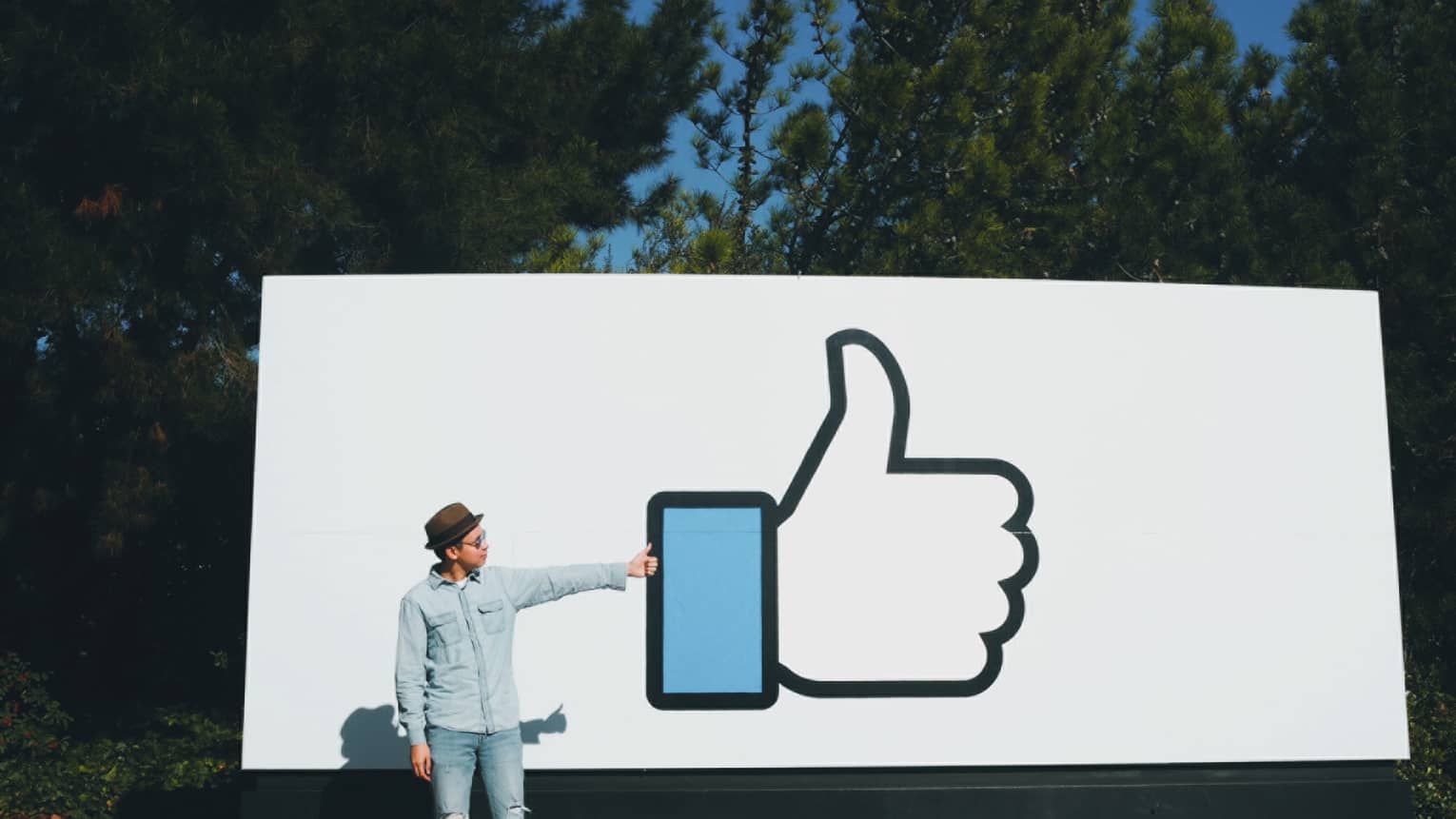 Man stands, gives thumbs-up in front of large white billboard sign with Facebook like icon