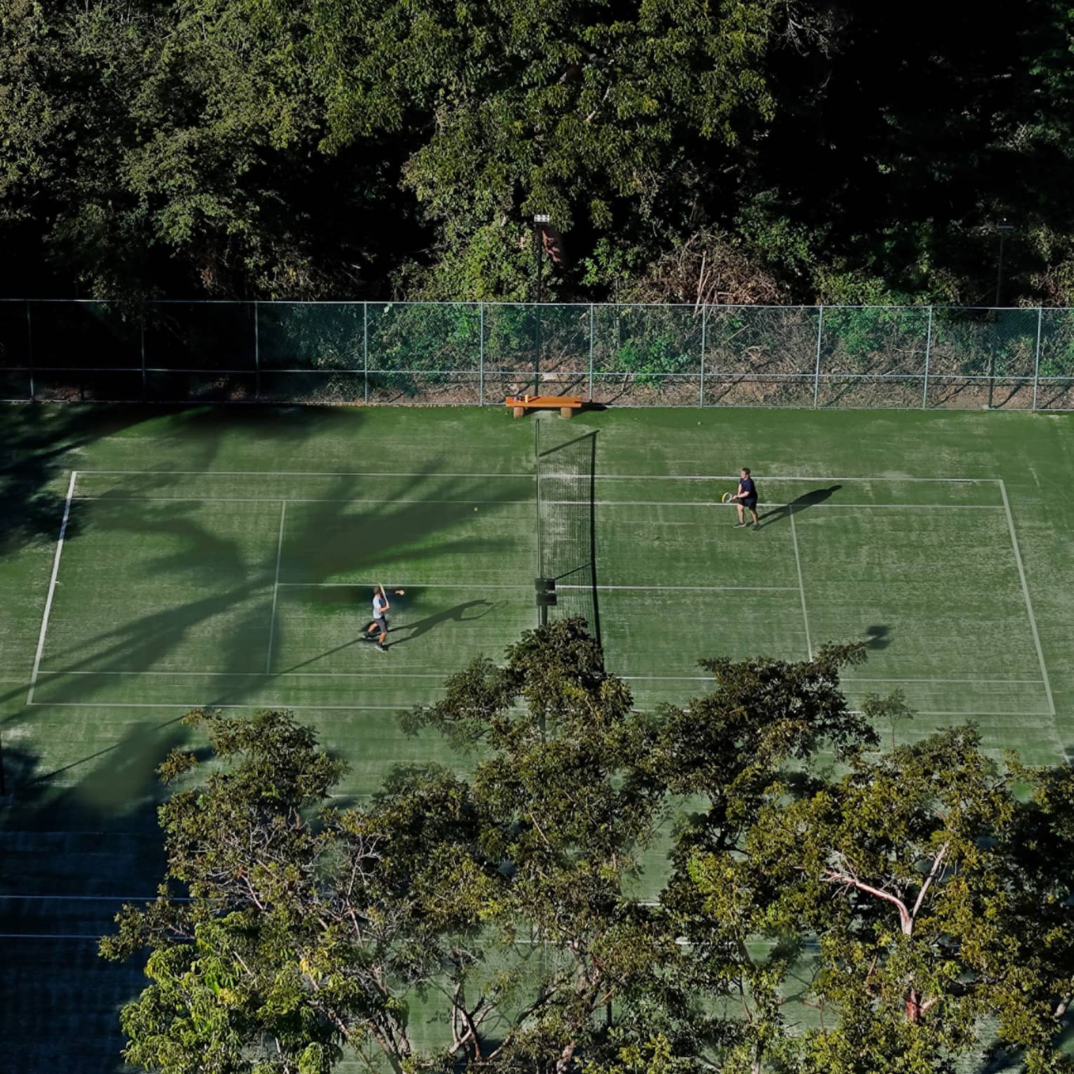 Two people play tennis on a green court surrounded by trees.