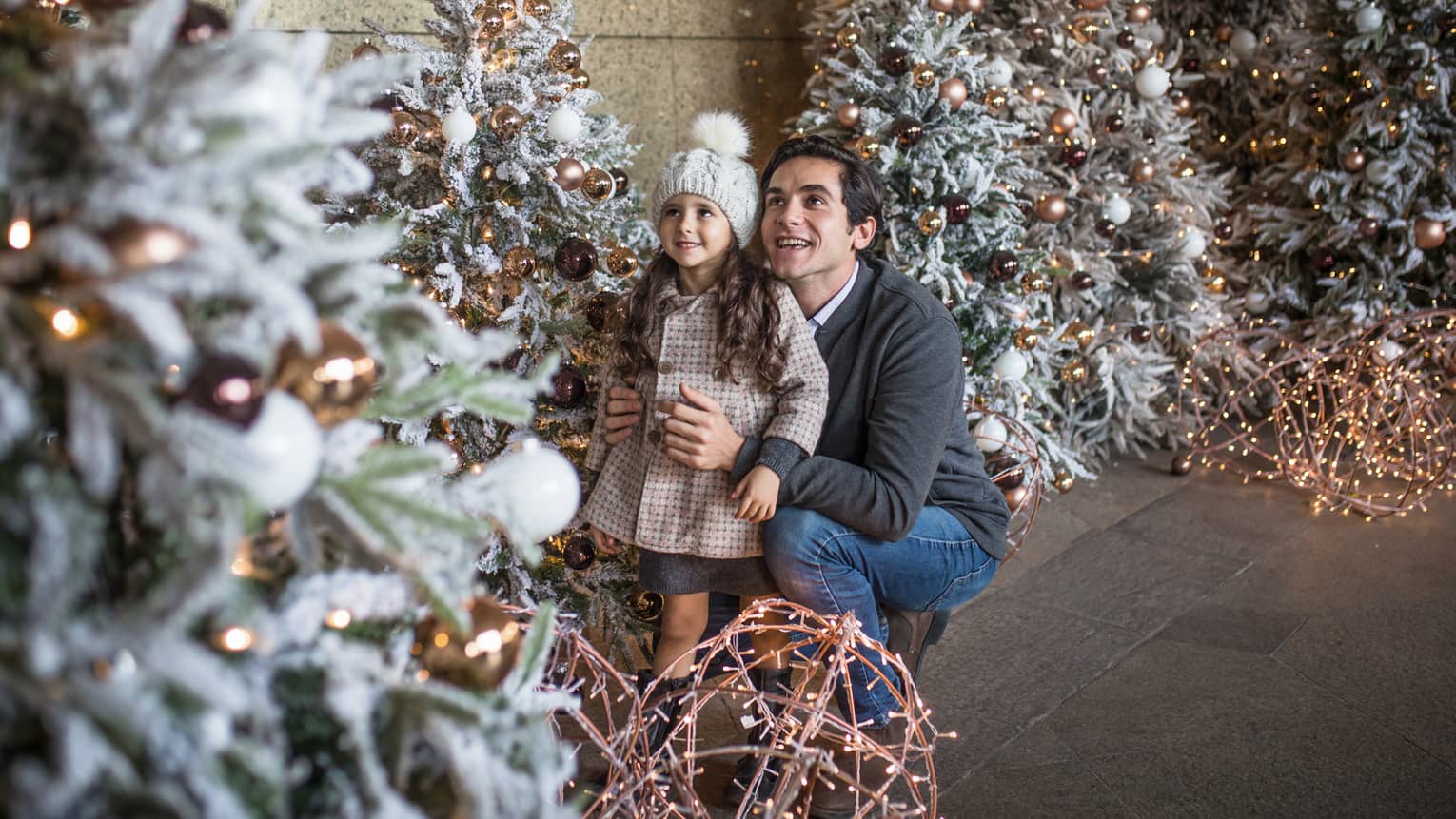 Man crouched with arms around girl in coat and hat admiring Christmas tree