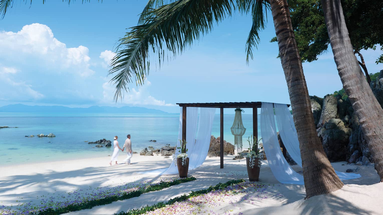 Couple walks on white sand beach near cabana with white curtains, large palm trees