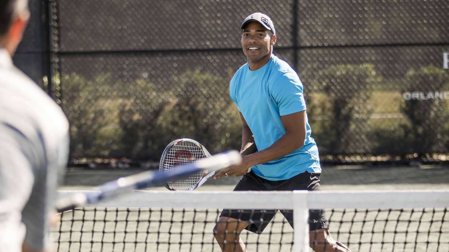 Man wearing blue T-shirt and hat holds racket as tennis ball flies over net