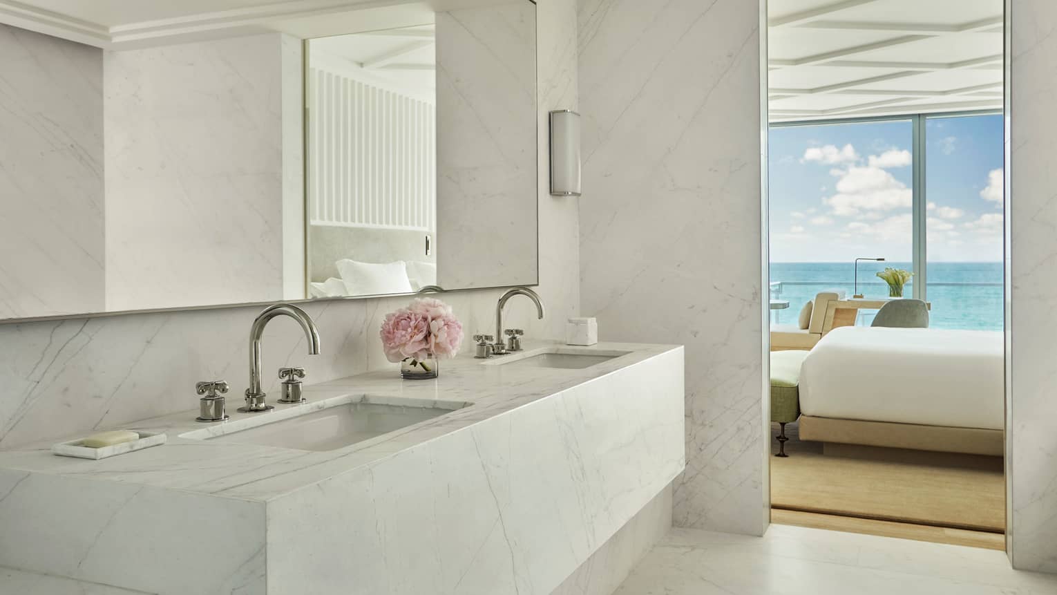 Luxury hotel bathroom with a marble double sink vanity, a mirror and a view into the bedroom with ocean views through floor-to-ceiling windows