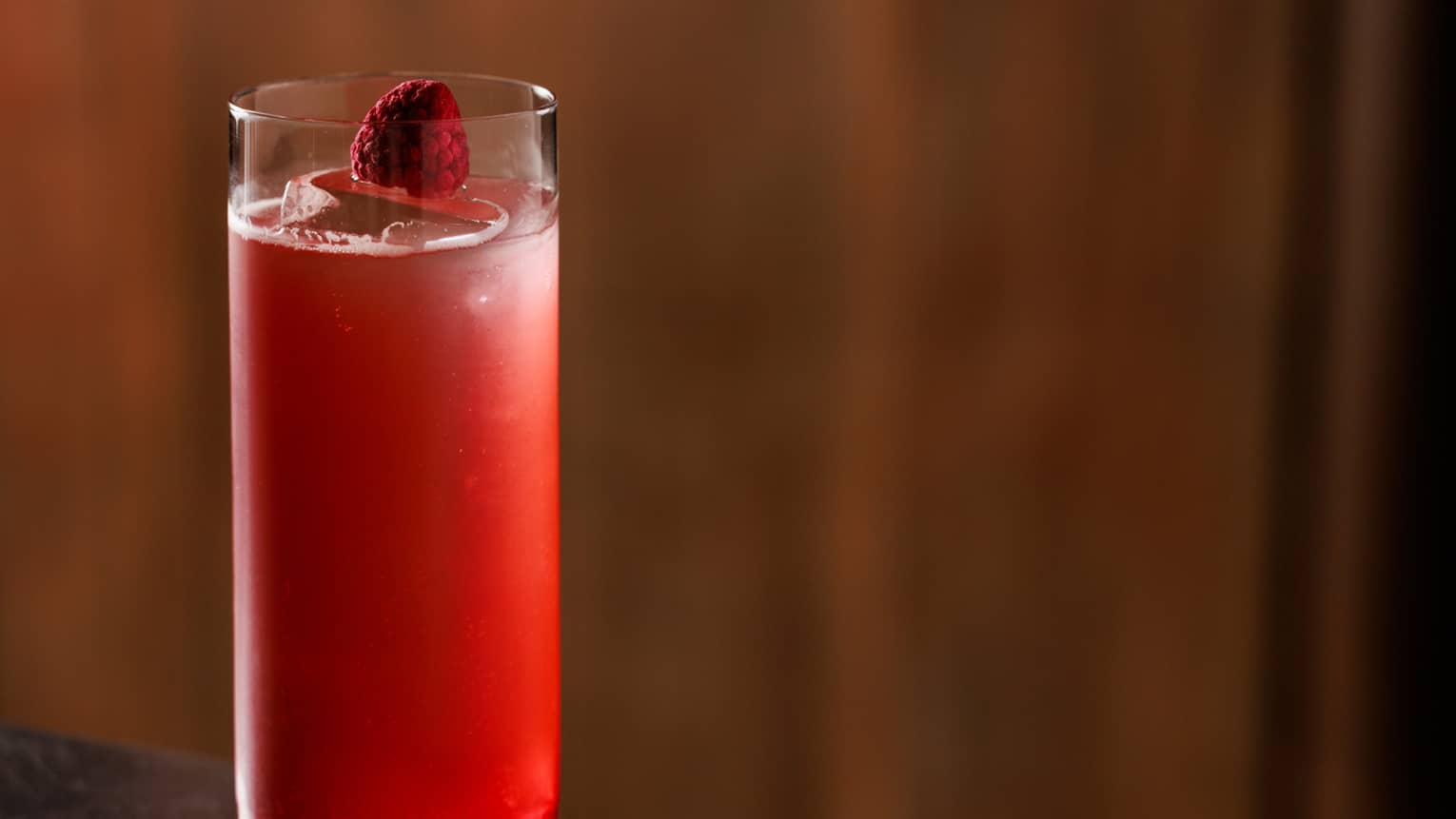 Red-coloured cocktail garnished with a single raspberry served in a highball glass