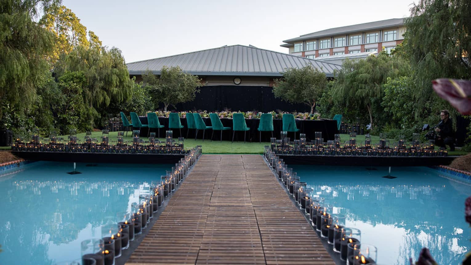 Wooden over-water walkway lined with candles in hurricane vases leads up to a platform where a large dining table set for a reception sits in the centre