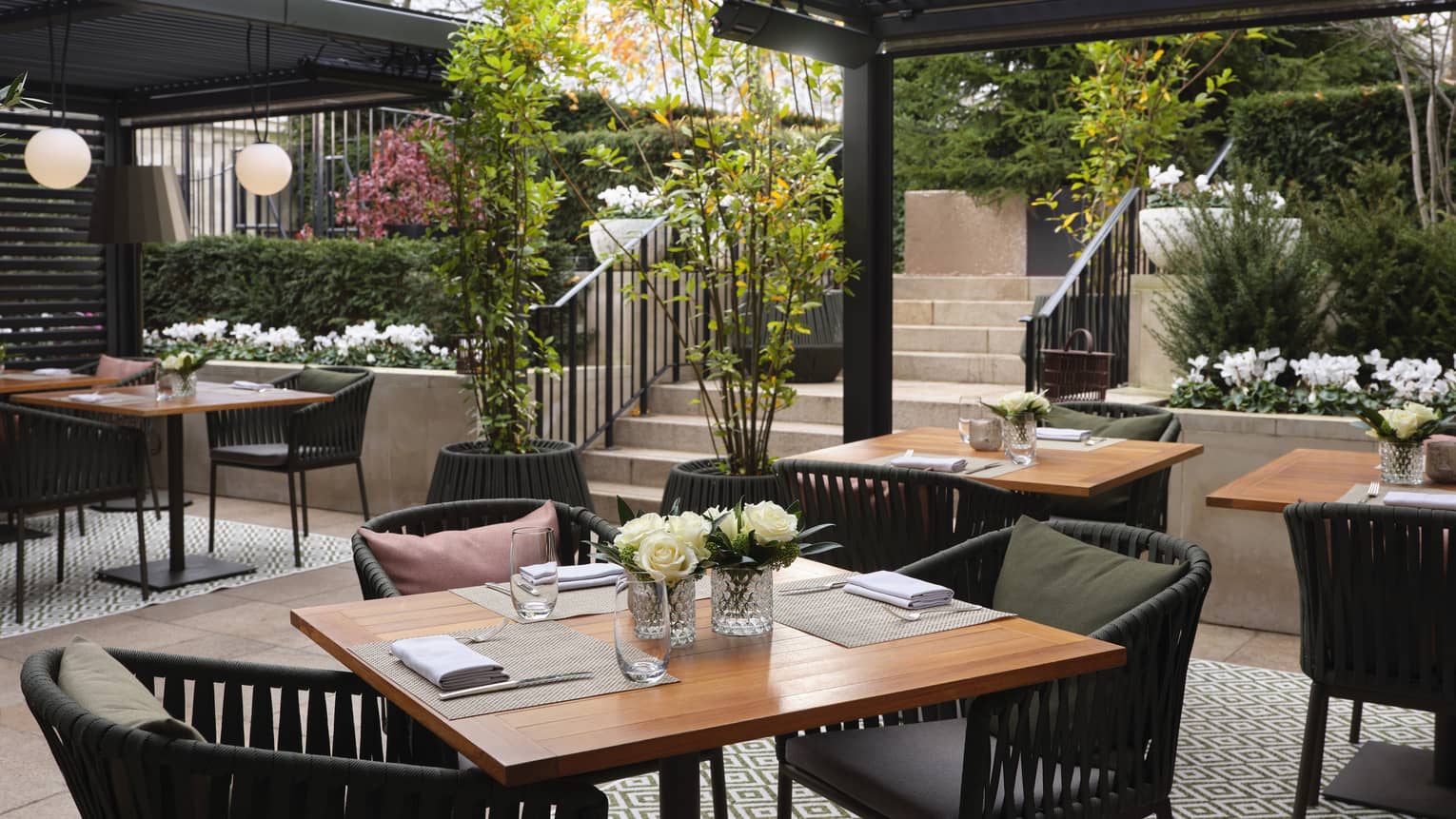 Set square wooden tables, dark rattan chairs, outdoor diamond-patterned rug under pergola