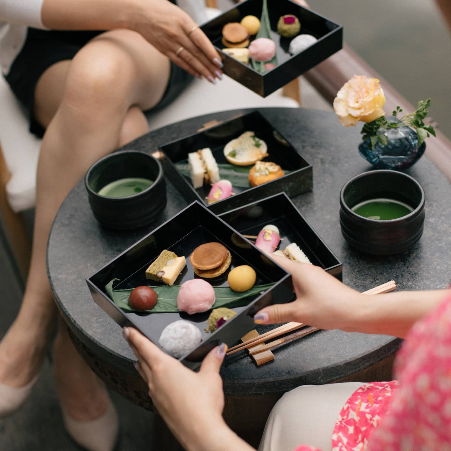 A small round table, with two-layered bento box of colourful, artfully arranged foods, top layers being lifted by two people.