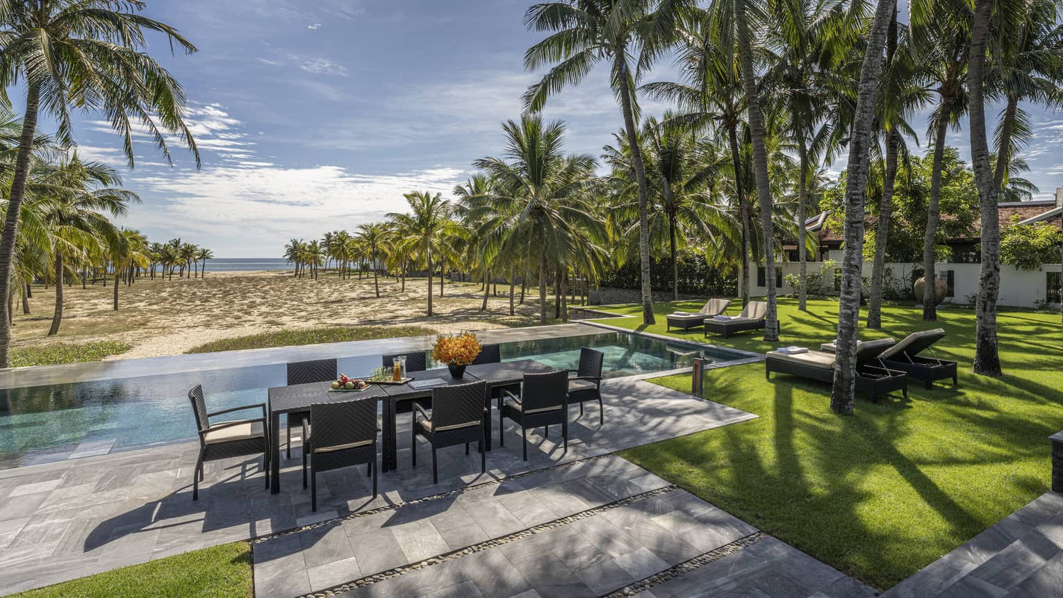 Four Bedroom Pool Villa pool area with dining table, lush green grass, palm trees and blue skies