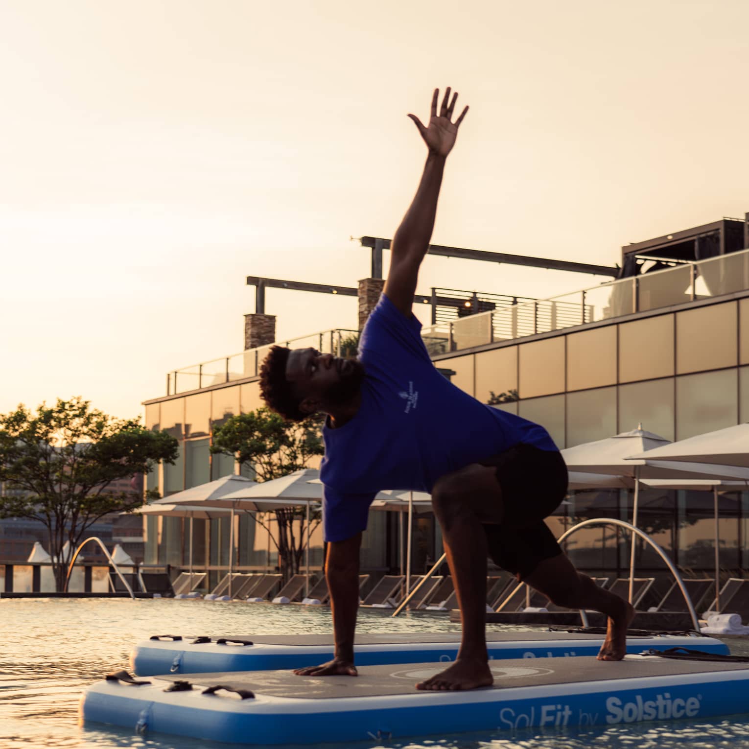 Side view of an instructor doing a yoga pose on a floating exercise board in a pool, one arm in the air, as the sun sets.