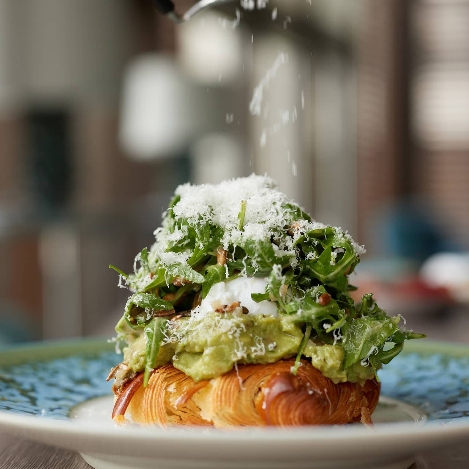 A close-up of a plate holding a croissant covered with avocado, egg and arugula salad topped with grated parmesan. 