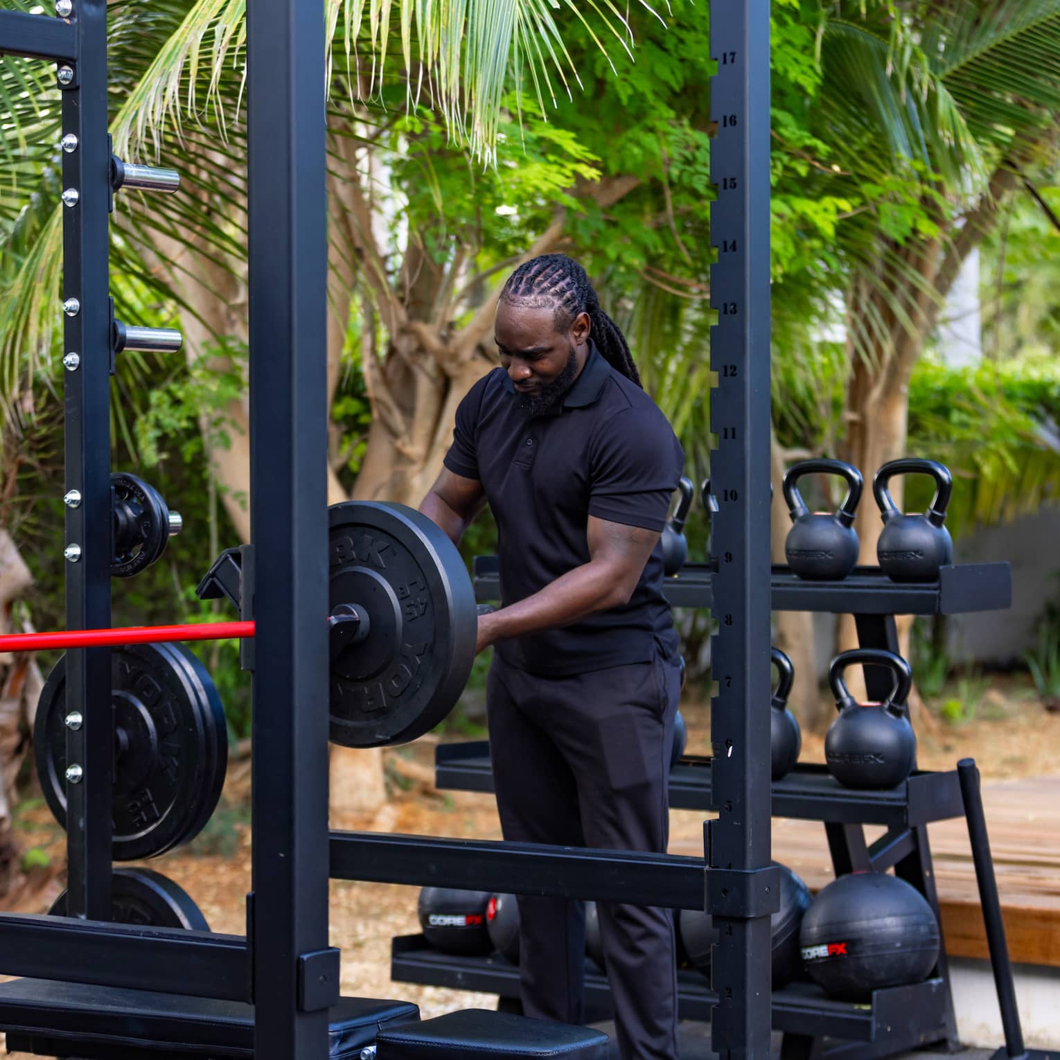 A guest adjusts a barbell in an outdoor gym against a rack of kettlebells and medicine balls and a grove of shady trees.