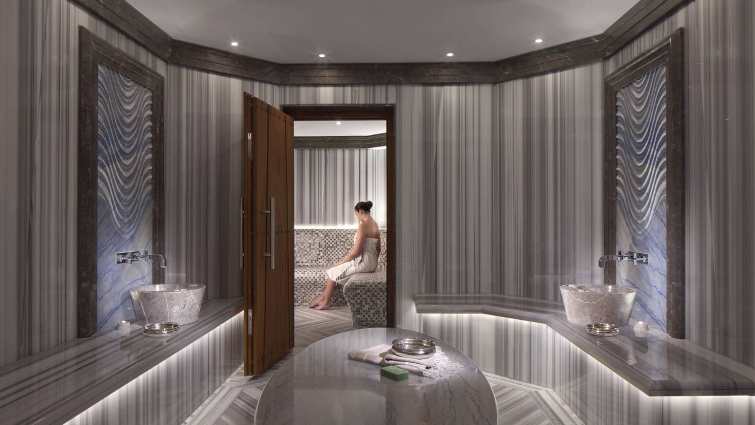 Elegant marble spa room with open wood door where woman wearing towel sits on bench 