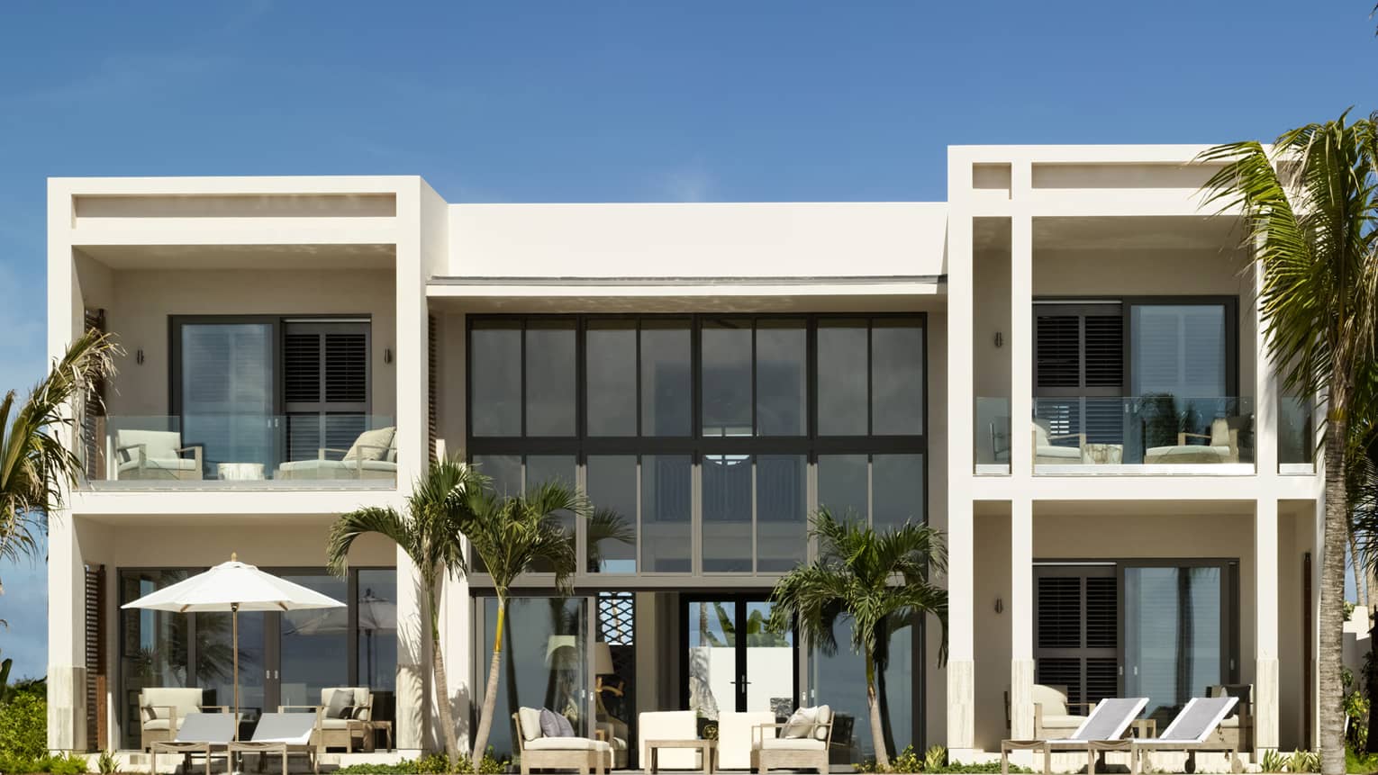 Luxury resort villa with large windows, private ocean-view balconies and lounge areas surrounded by tropical palm trees 