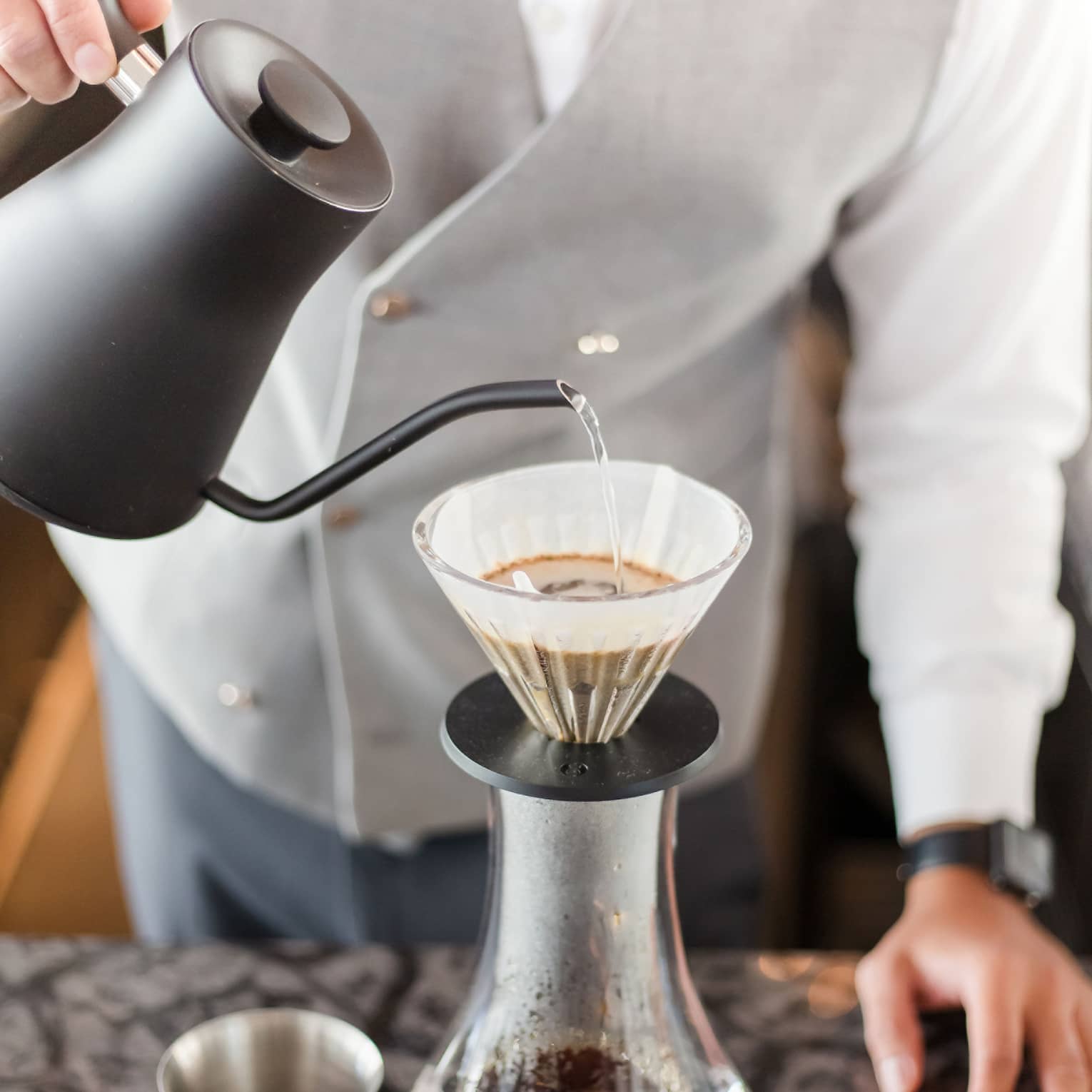 Barista holds black teapot, pours hot water over coffee in cone filter and glass jug on tray