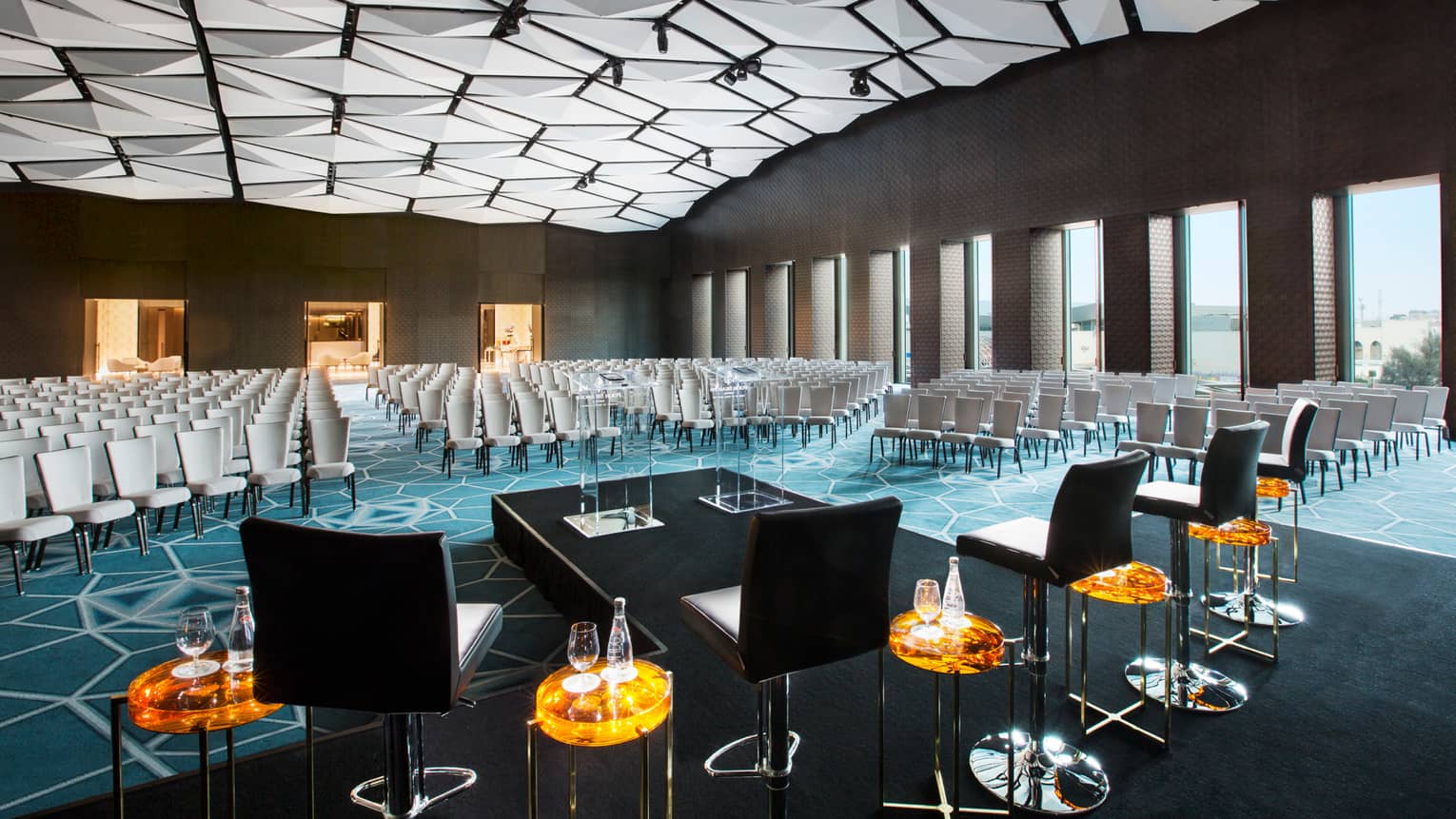 Rows of chairs fill large meeting room in front of stage, podium, geometric lights across ceiling