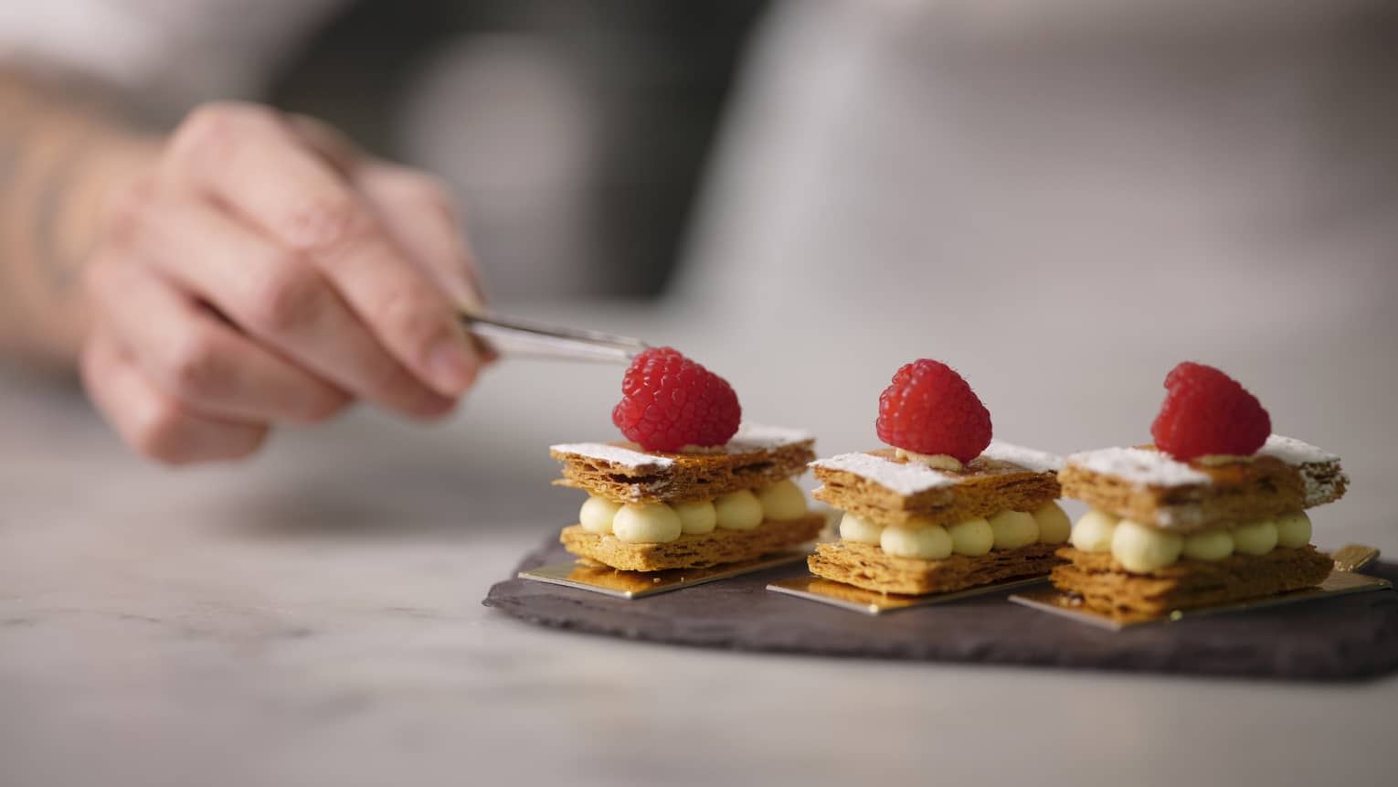 A chef placing a bit of sugar onto three scones topped with raspberries using small metal tongs