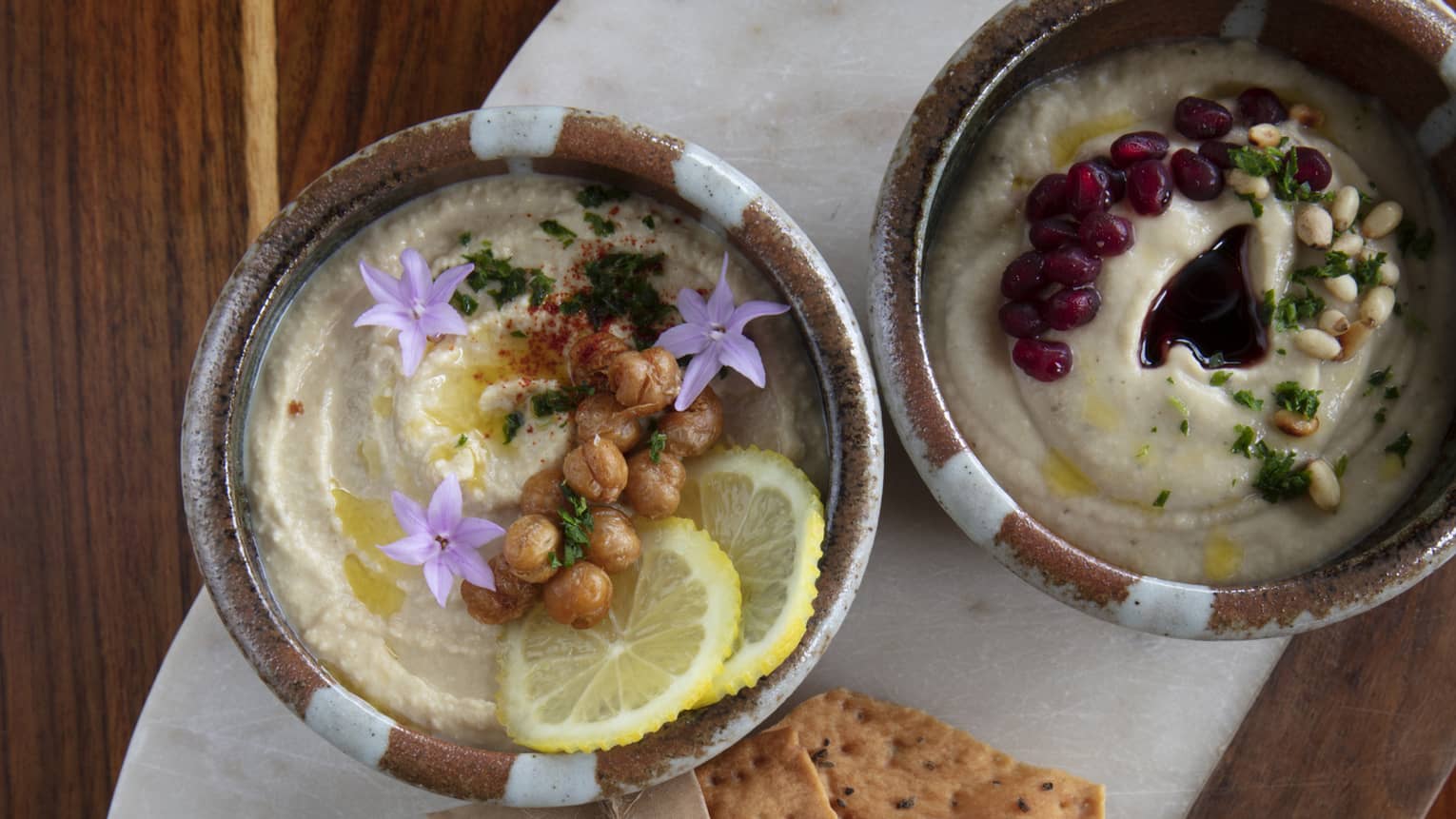 Aerial shot of roasted eggplant dip garnished with chickpeas, lemon and pomegranate seeds, accompanied by crackers on an oblong white marble slab