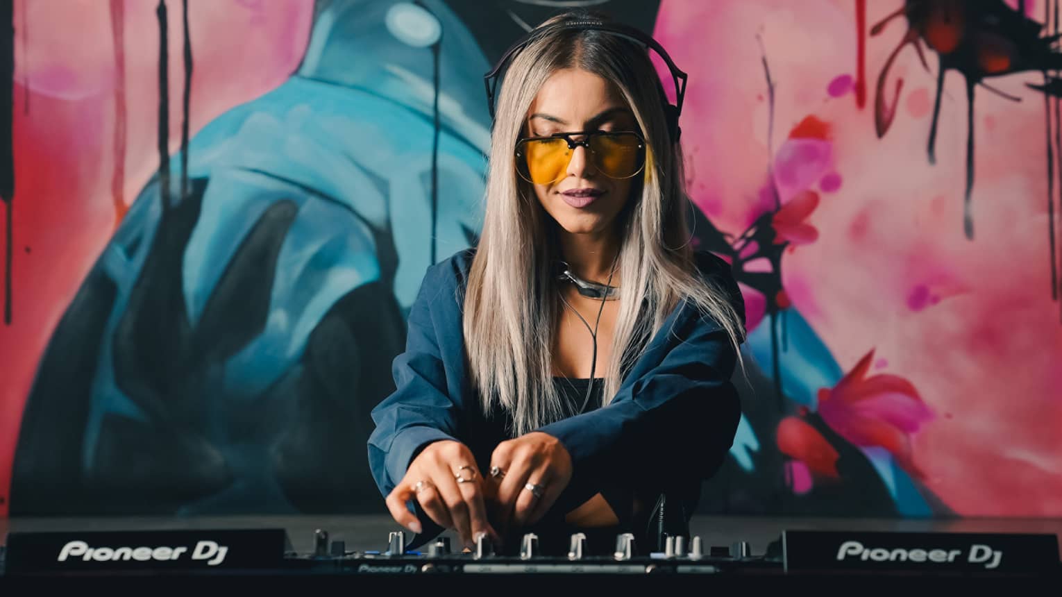 Female DJ with long blonde hair and orange-lensed sunglasses stands turning mixing knobs behind a DJ stand set up in front of a pink-and-blue mural of a woman