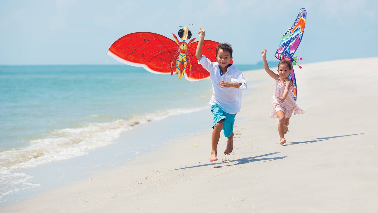 Two young children run with colourful butterfly kites on white sand beach