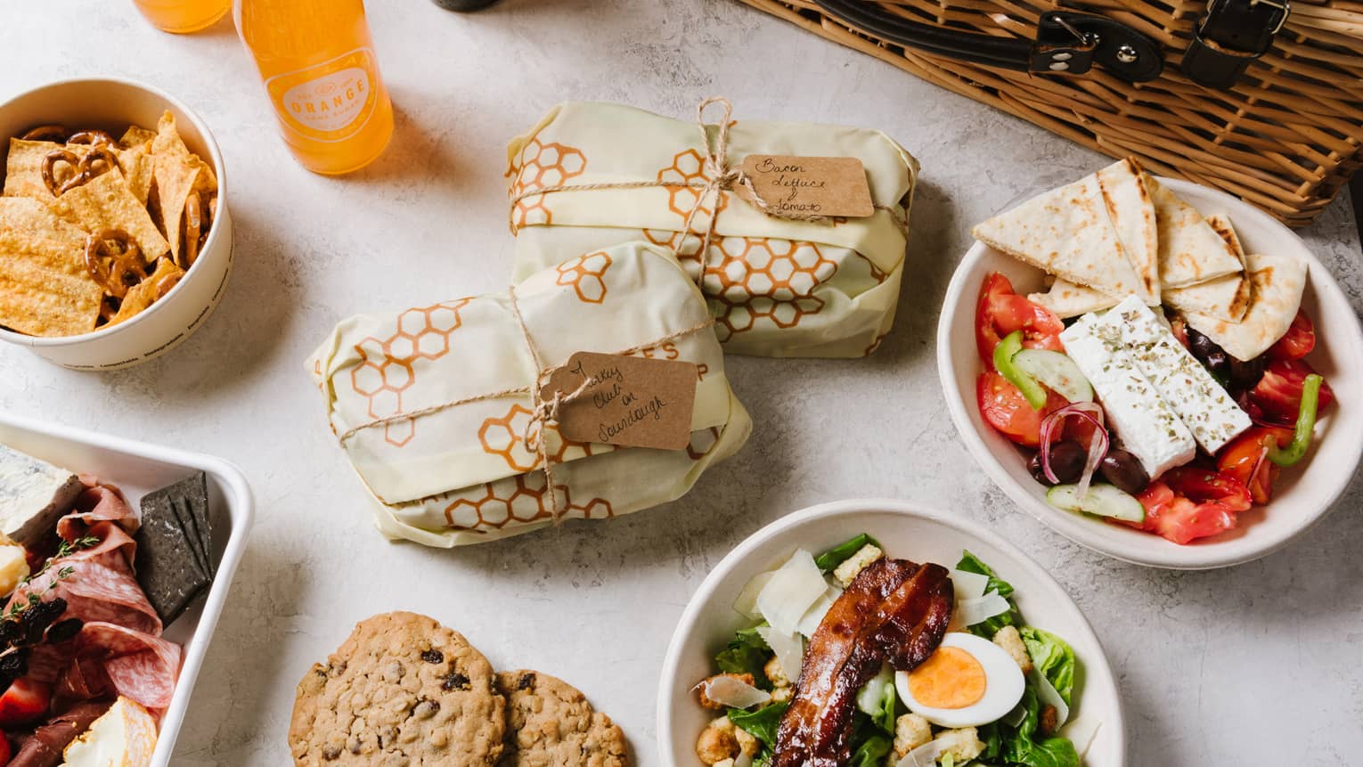 An assortment of picnic food including salad, cookies, sandwiches, fruit, and sliced meat next to a brown wood basket.