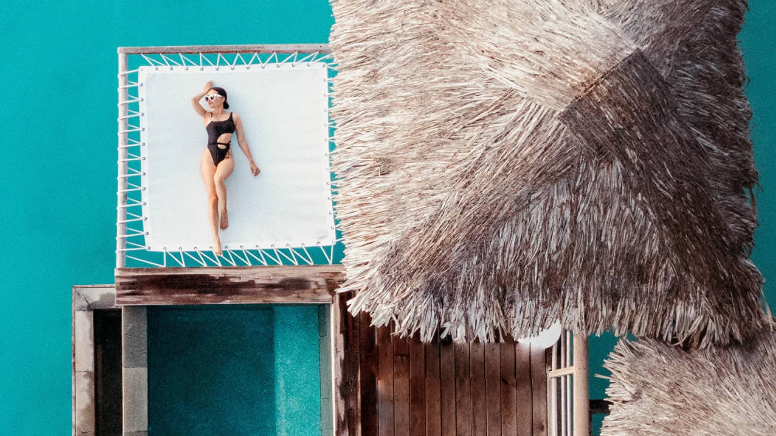 Woman lays on overwater net next to thatched-roof bungalow