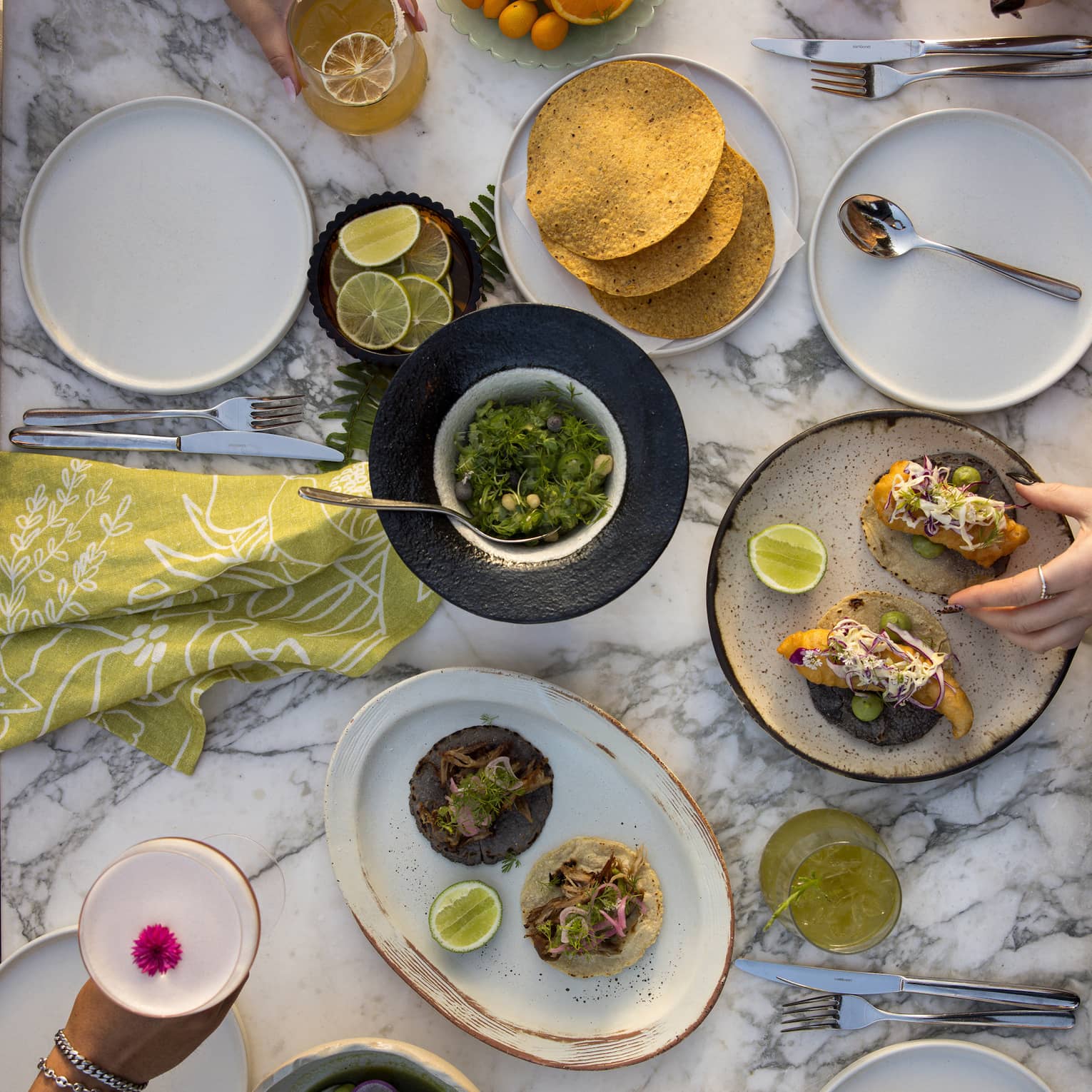 Overhead view of white marble table filled with plates of tacos, bowls of salsa, slices of oranges and limes, with two hands holding cocktails and another set of hands reaching for food