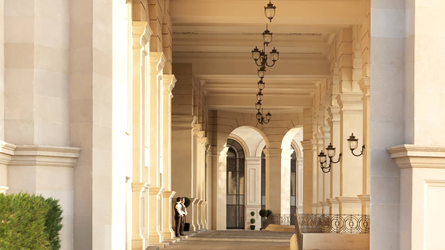 Two doormen stand side-by-side in front of hotel, exterior building with row of black lanterns above