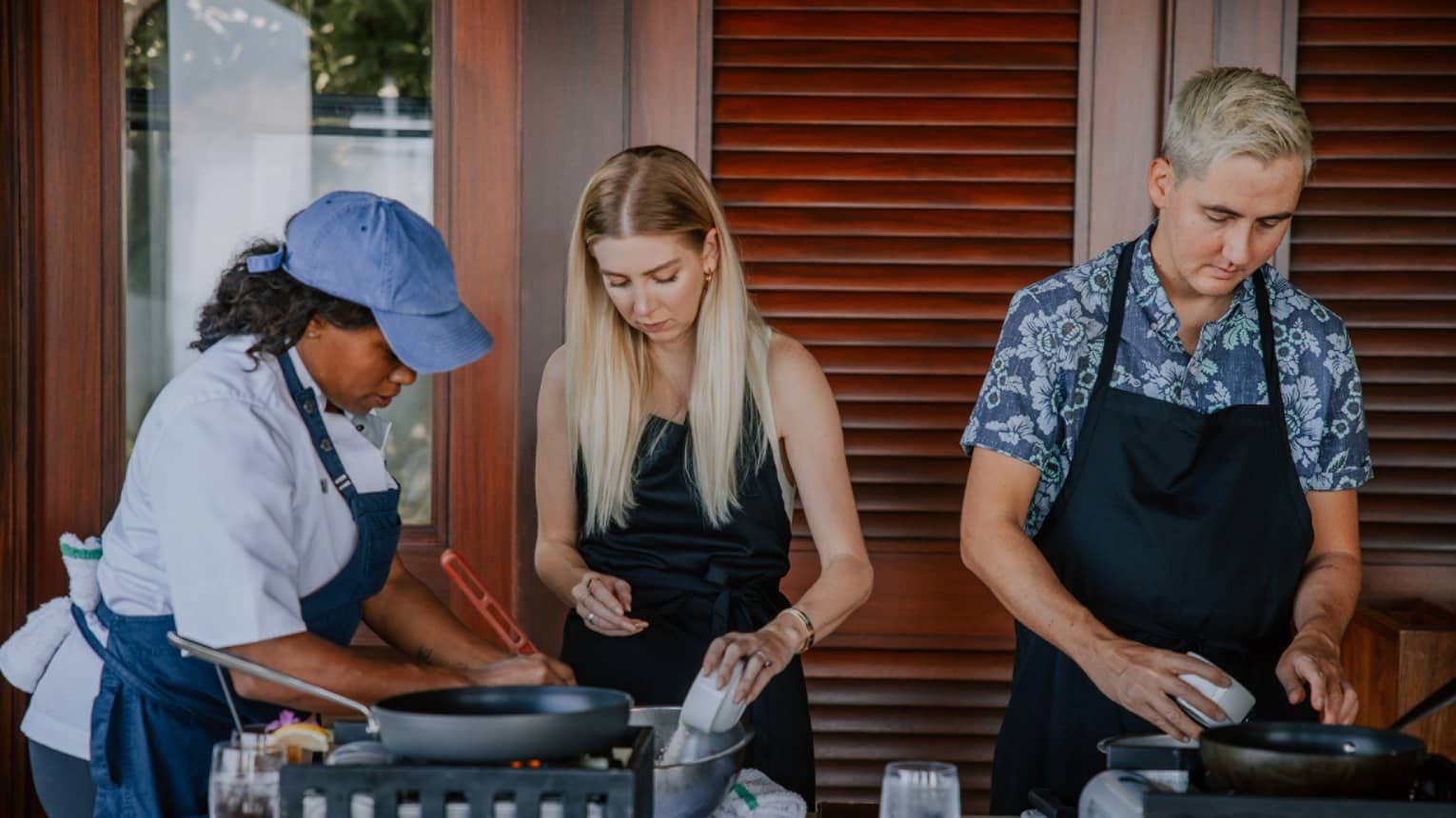 A cooking class is underway as three people sporting aprons stand over a table strewn with wine glasses and cookware.