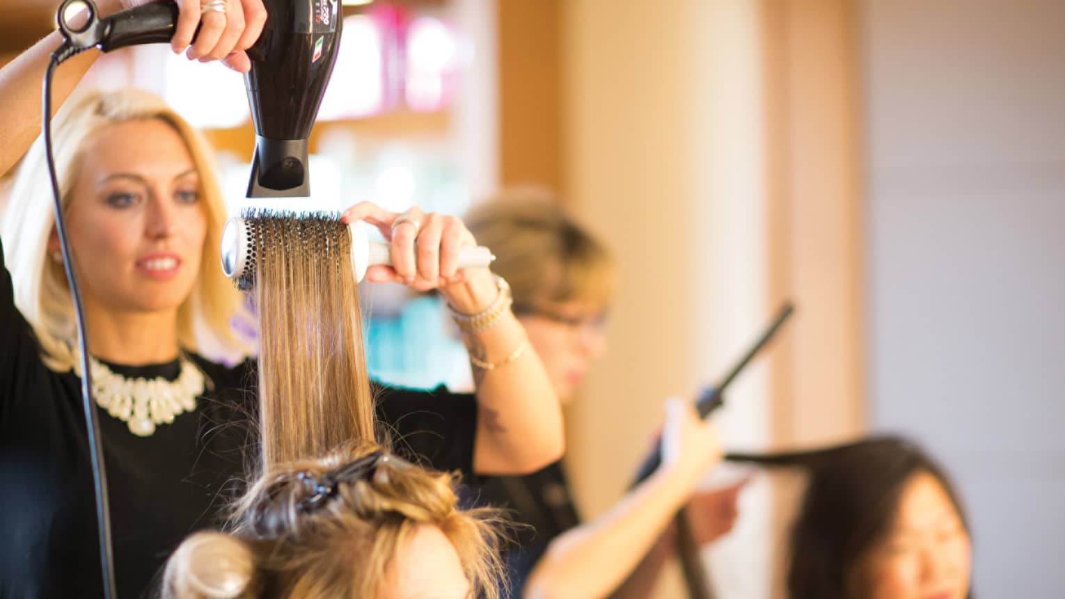 Hairstylist with hairdryer pulls back woman's hair with round brush in salon