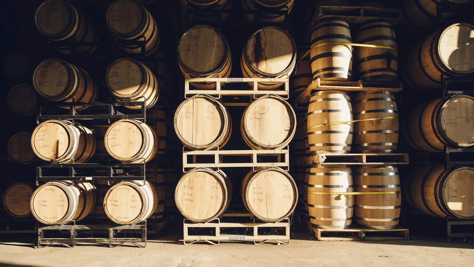 Several wooden barrels lined up on racks in a distillery with information about what is in each barrel written on top.