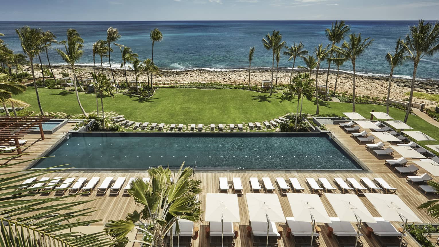 Aerial view of large outdoor swimming pool lined with white deck chairs, lawn, beach