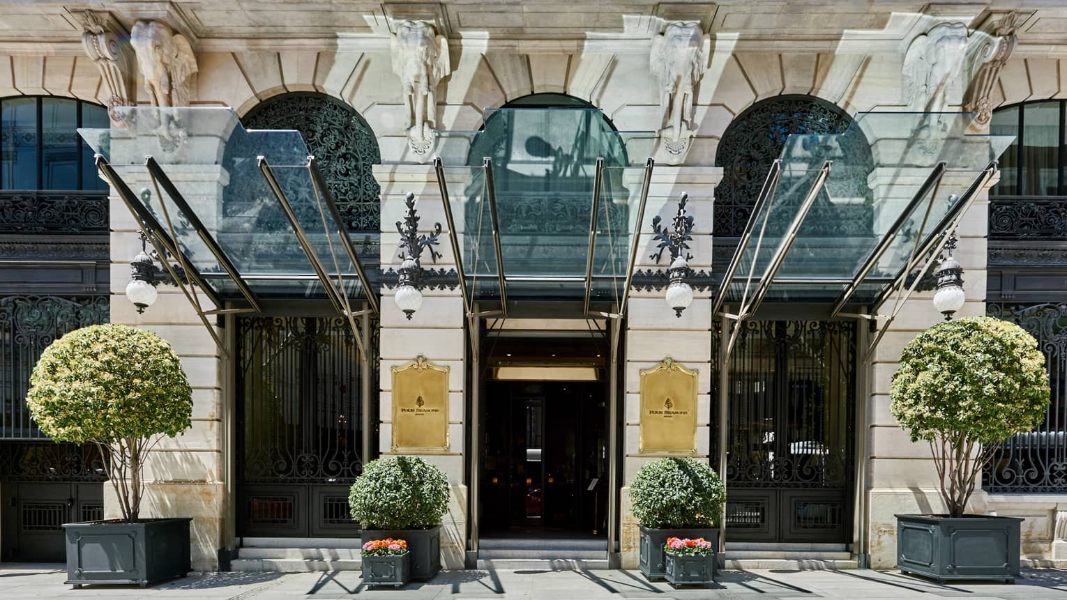 Exterior view of Hotel with potted foliage, glass canopy and golden plaques, second-story balcony visible