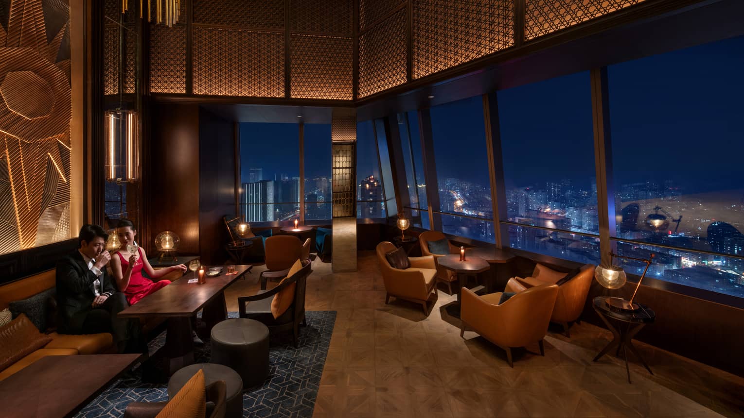 A couple enjoys drinks in a darkly lit bar with ornate lighted ceiling panelling and an aerial view of the city at night.